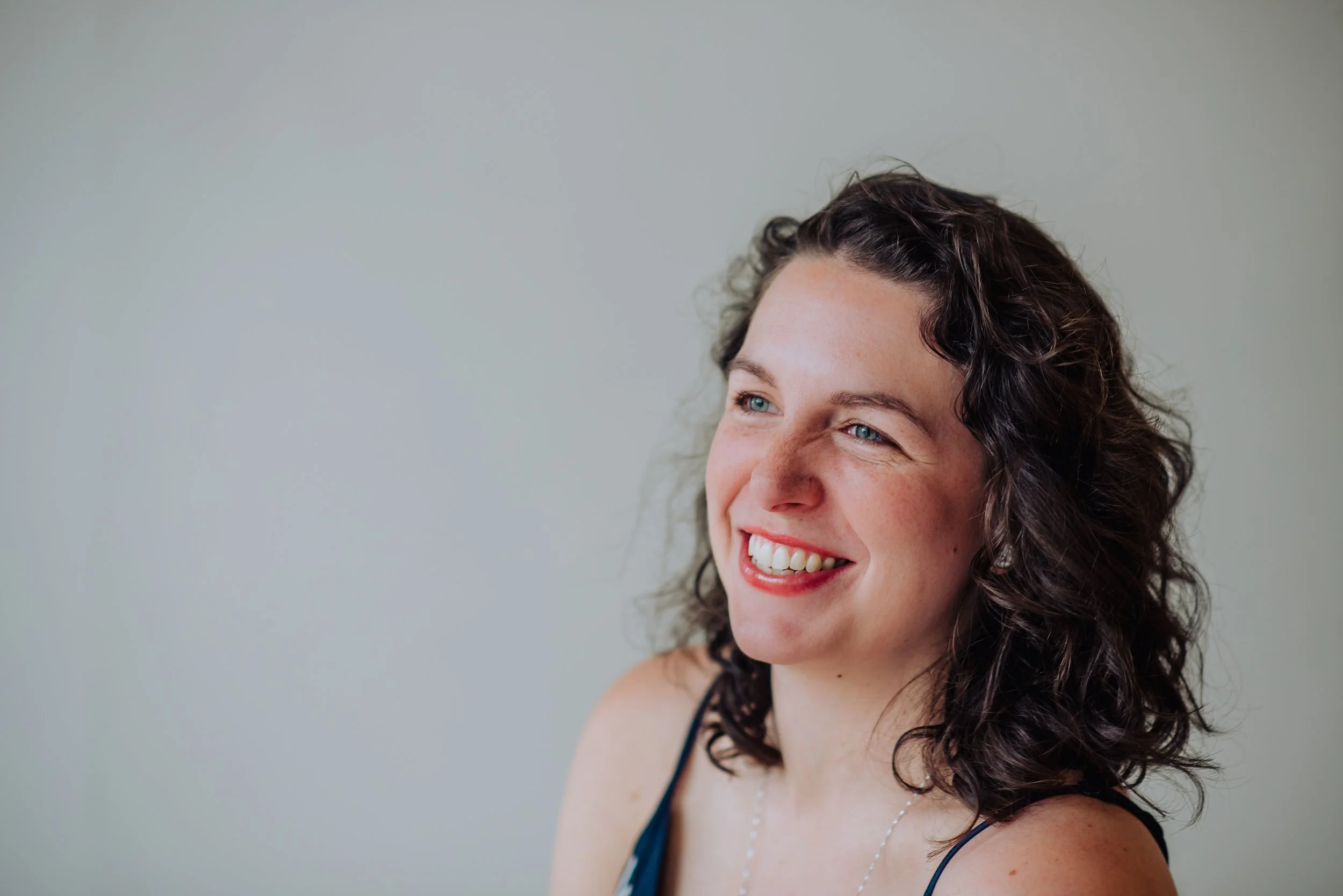 A woman with curly dark hair and bright blue eyes smiling, wearing a black spaghetti strap top and a silver necklace.