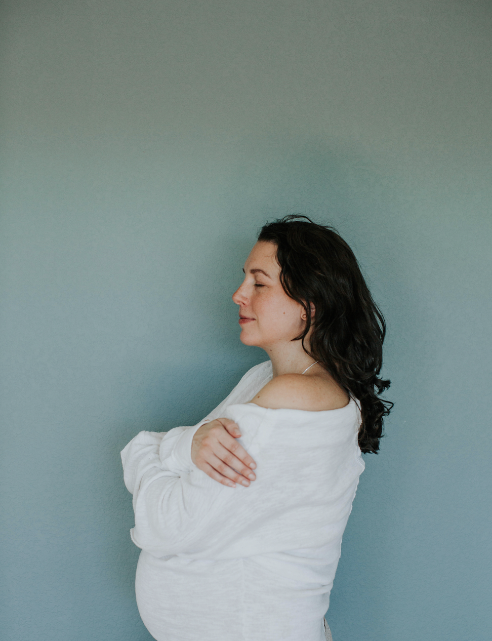 Pregnant woman with dark curly hair, wearing a white top, standing with arms crossed, smiling softly against a light blue wall.