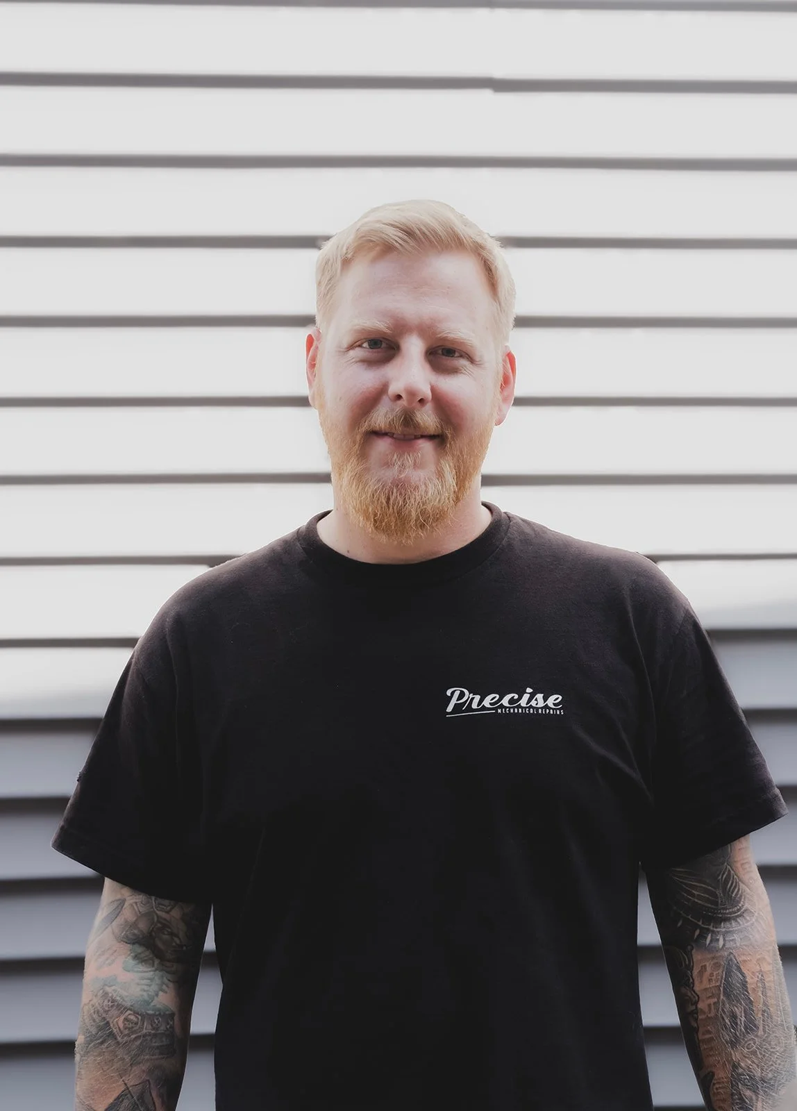 A man with a beard and short blonde hair smiling in a workshop. He is wearing a navy blue work uniform with a name tag that reads "Adam" and another that says "Precise". Behind him, there are hooks with hanging dark blue jeans, shelves with tools, and storage boxes.