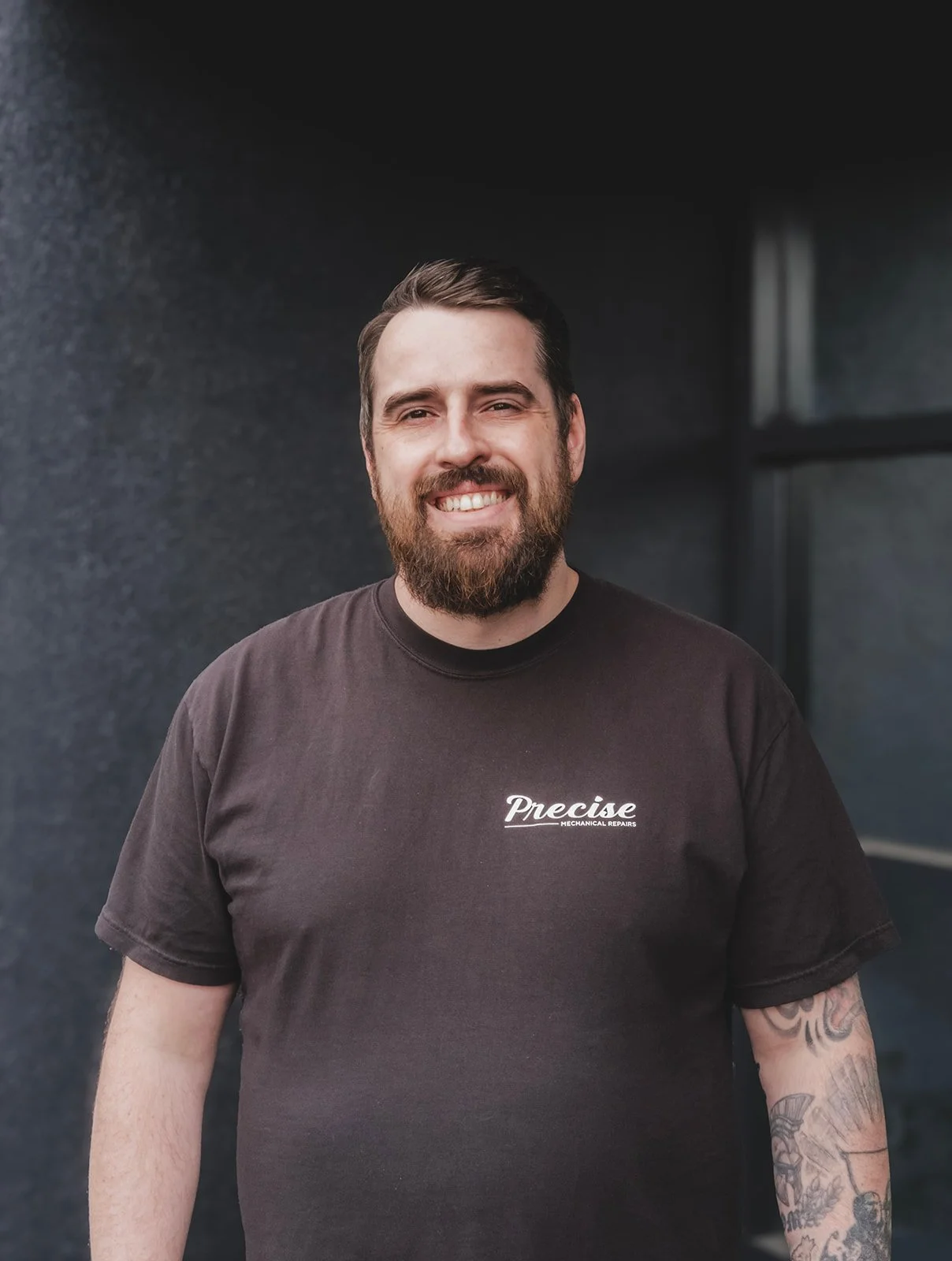 A man with a beard and tattoos, wearing a black t-shirt with the logo 'Precise Mechanical Repairs', smiling in front of a dark wall and window.