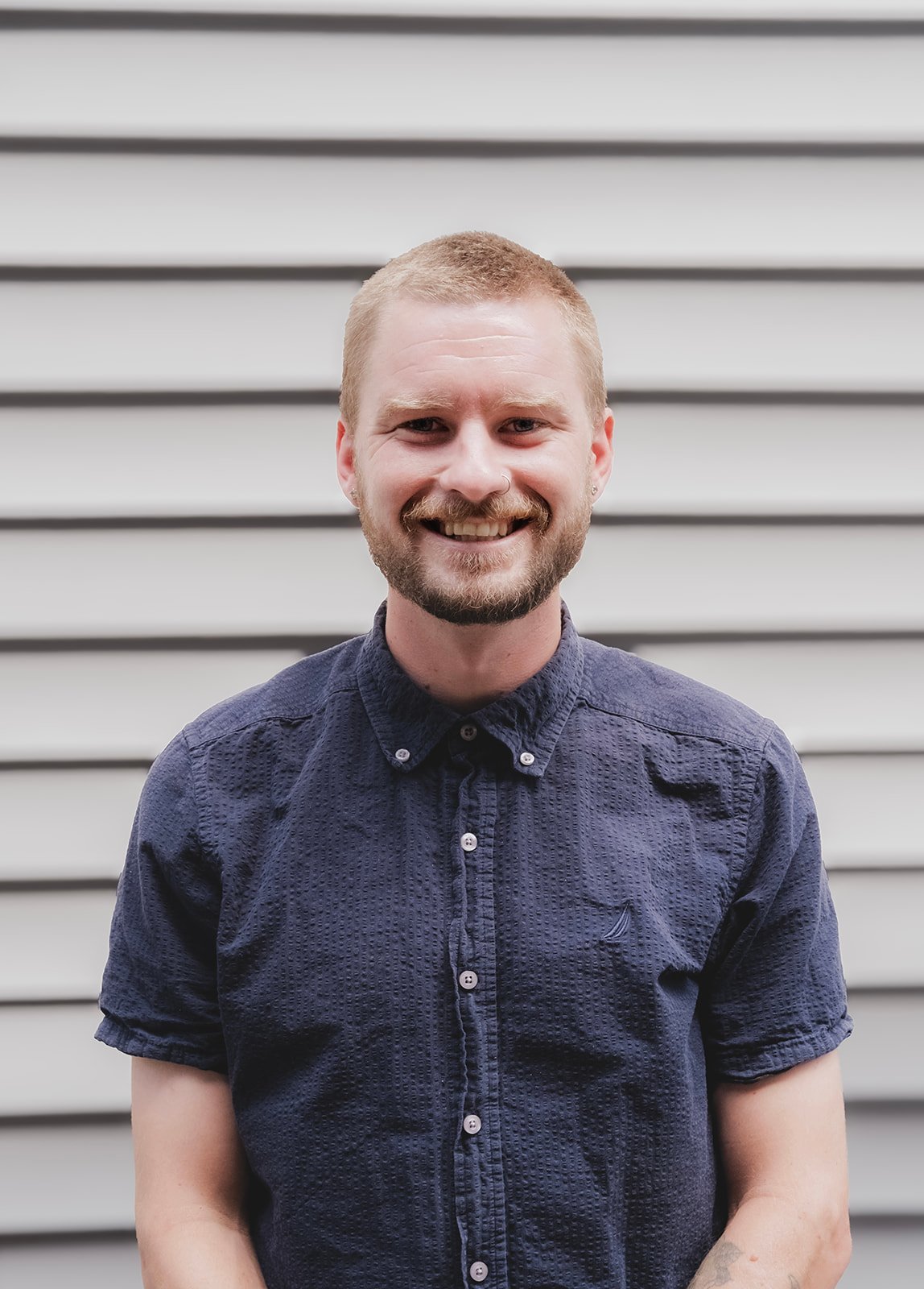 A young man with light brown hair, a nose ring, and a beard, smiling in front of a sign that reads 'Reclat Mechanical' inside a room with dark green walls.