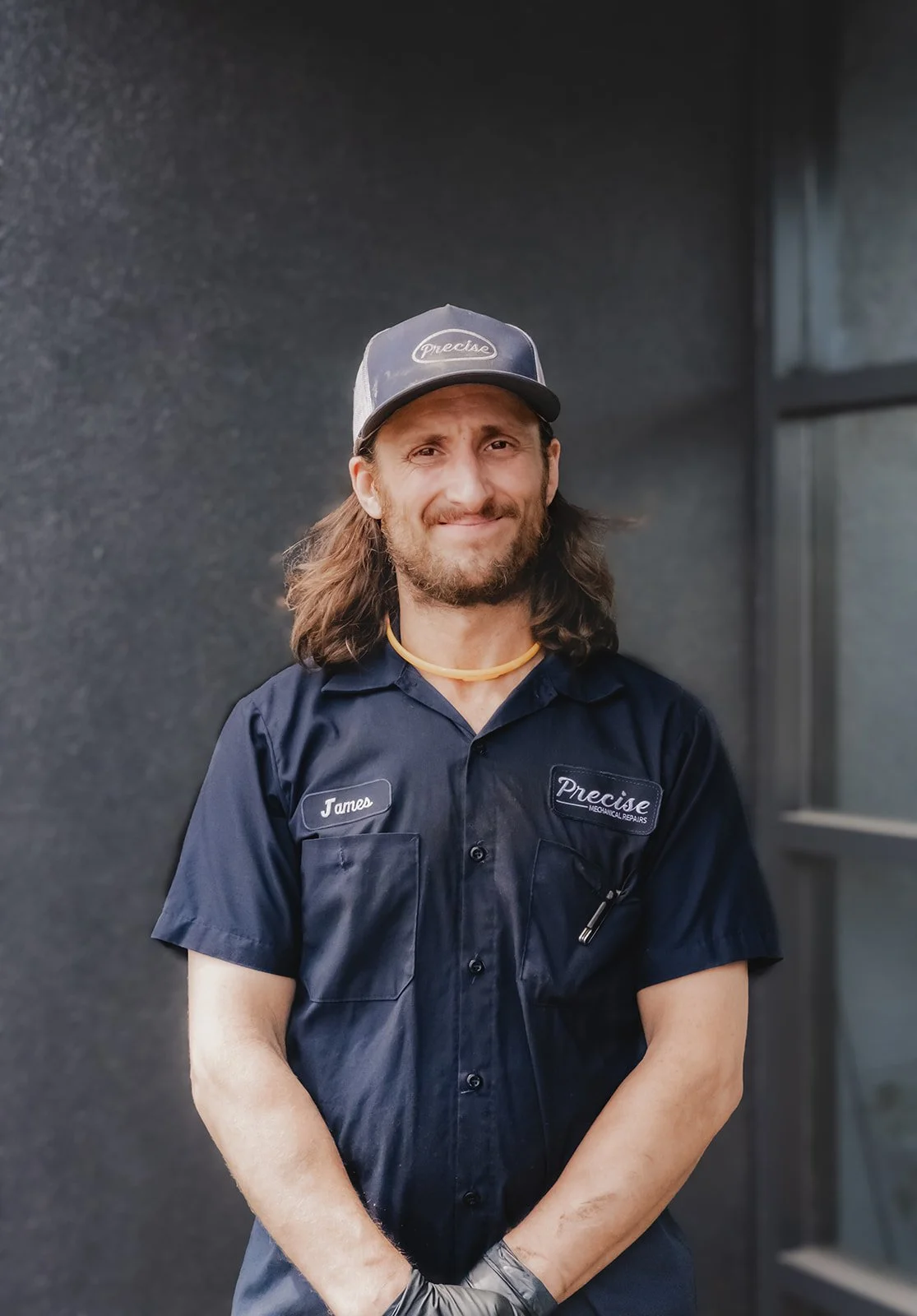 A man with a beard and wavy hair, wearing a navy blue mechanics jumpsuit with a name tag reading 'James' and a patch that says 'Precise Mechanical Repairs', standing in a garage or workshop.
