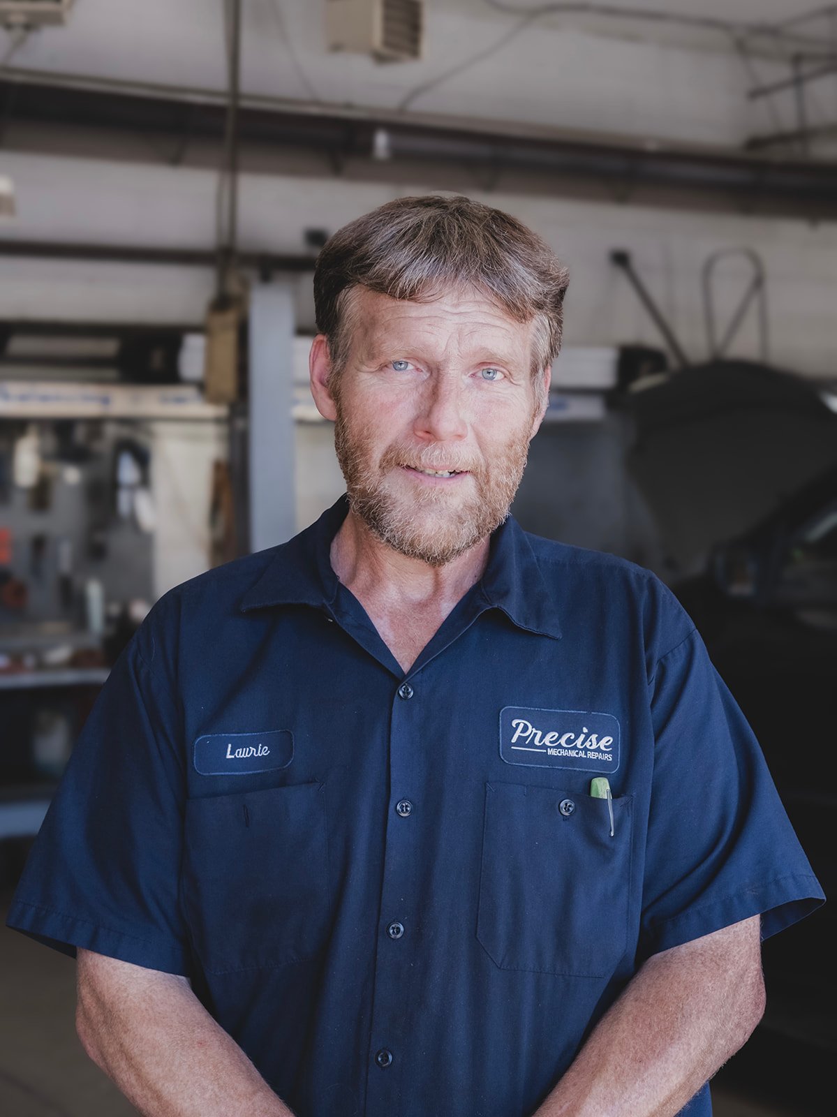 A man with a beard, wearing a blue work uniform with the name tag 'Laurie' and a patch that reads 'Precise Mechanics'