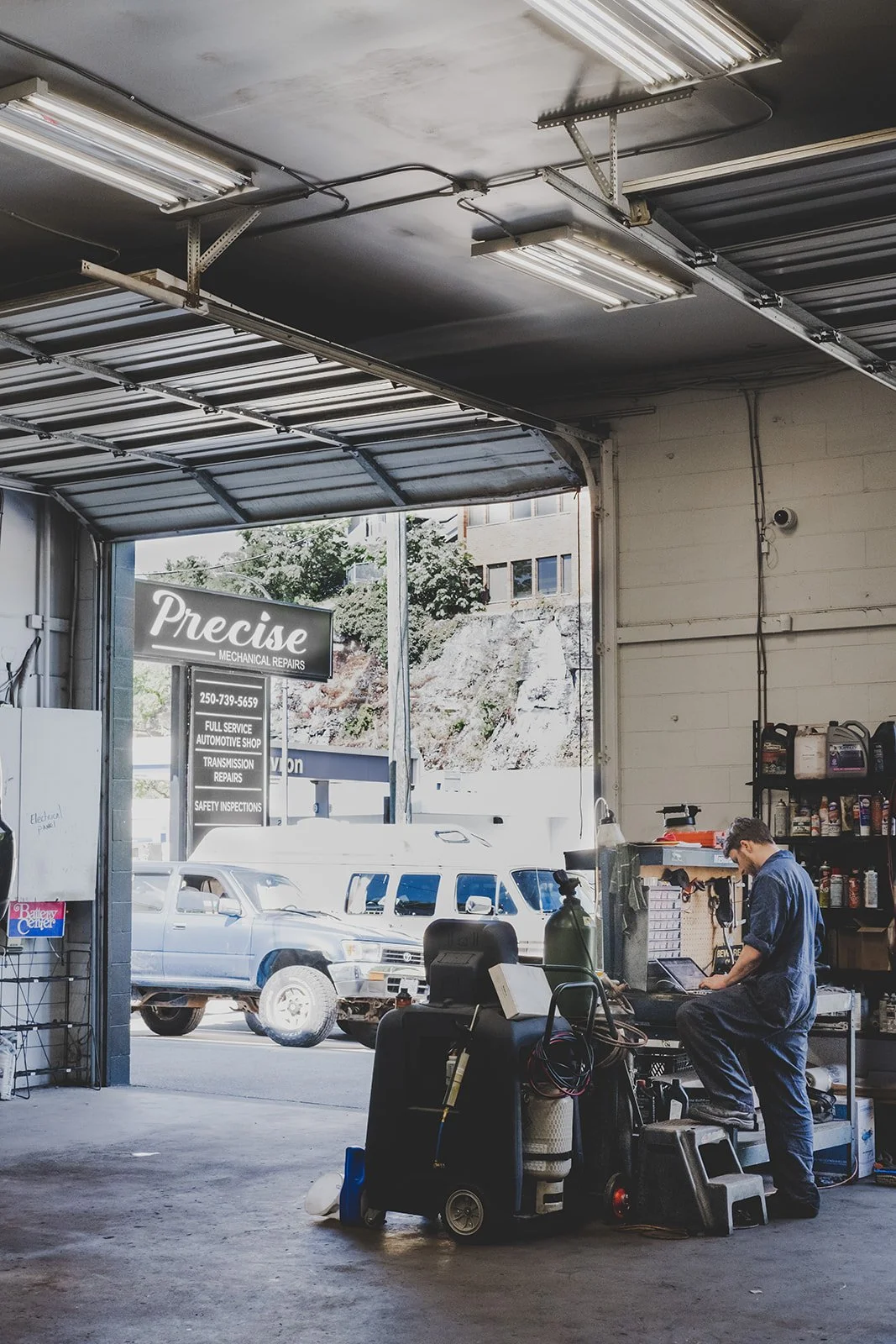 Inside an auto repair shop with a mechanic working on a laptop, shelves with car supplies, and cars parked outside visible through the open garage door.