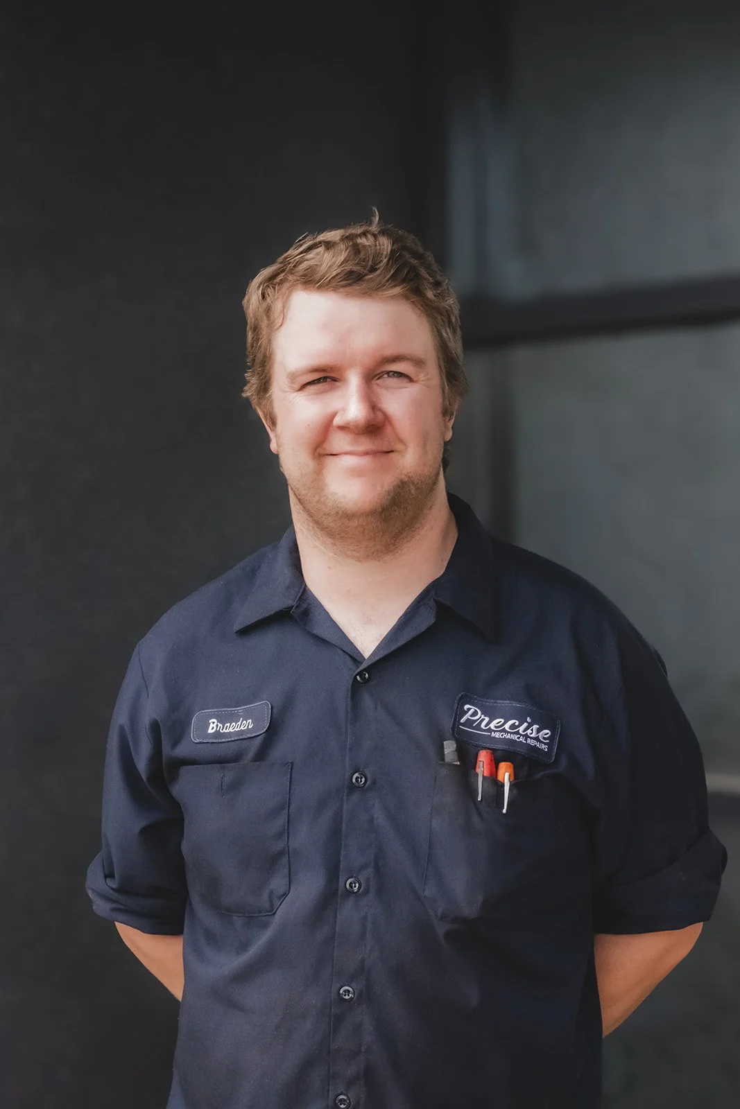 A man with light brown hair and a slight smile, wearing a dark blue work shirt with name tags that read 'Brenden' and 'Precise Mechanical Repairs,' stands outdoors against a dark, modern building wall background.