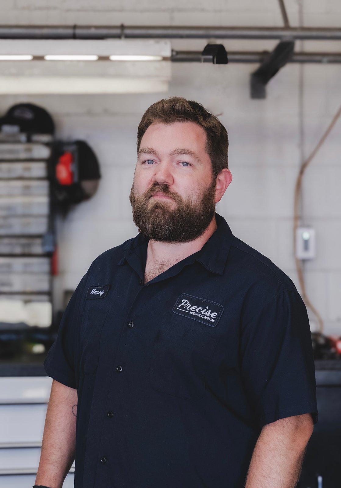 A man with a beard wearing a black mechanic shirt with patches reading 'Harry' and 'Precise Mechanical Repair', standing in a workshop with tools and equipment in the background.