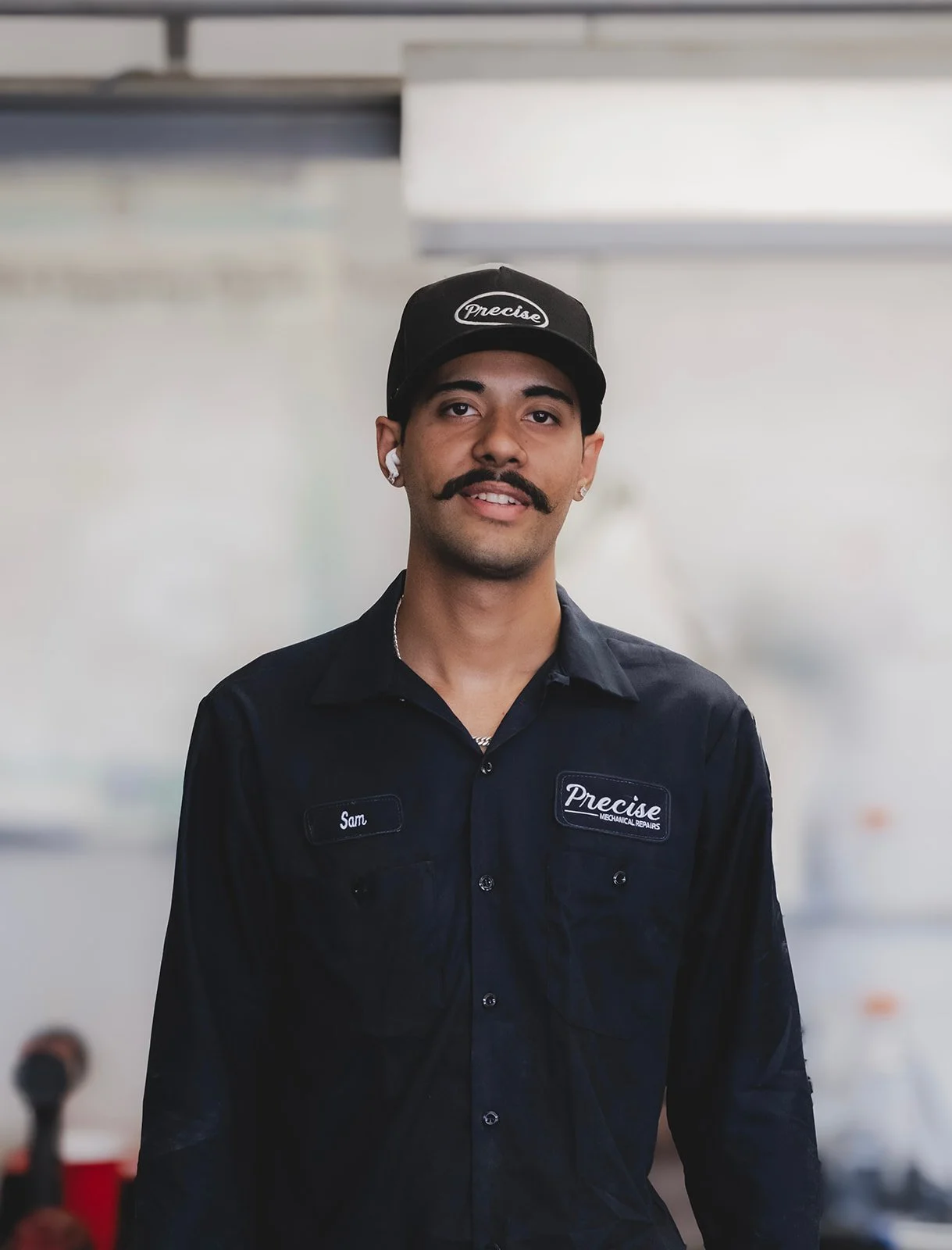 A man with a mustache and short hair, wearing a black Cap with 'Precise' embroidered on it, and a dark work shirt with a name tag that says 'Sam' and 'Precise Mechanical Repairs.' He is smiling slightly and standing indoors with a blurred background.