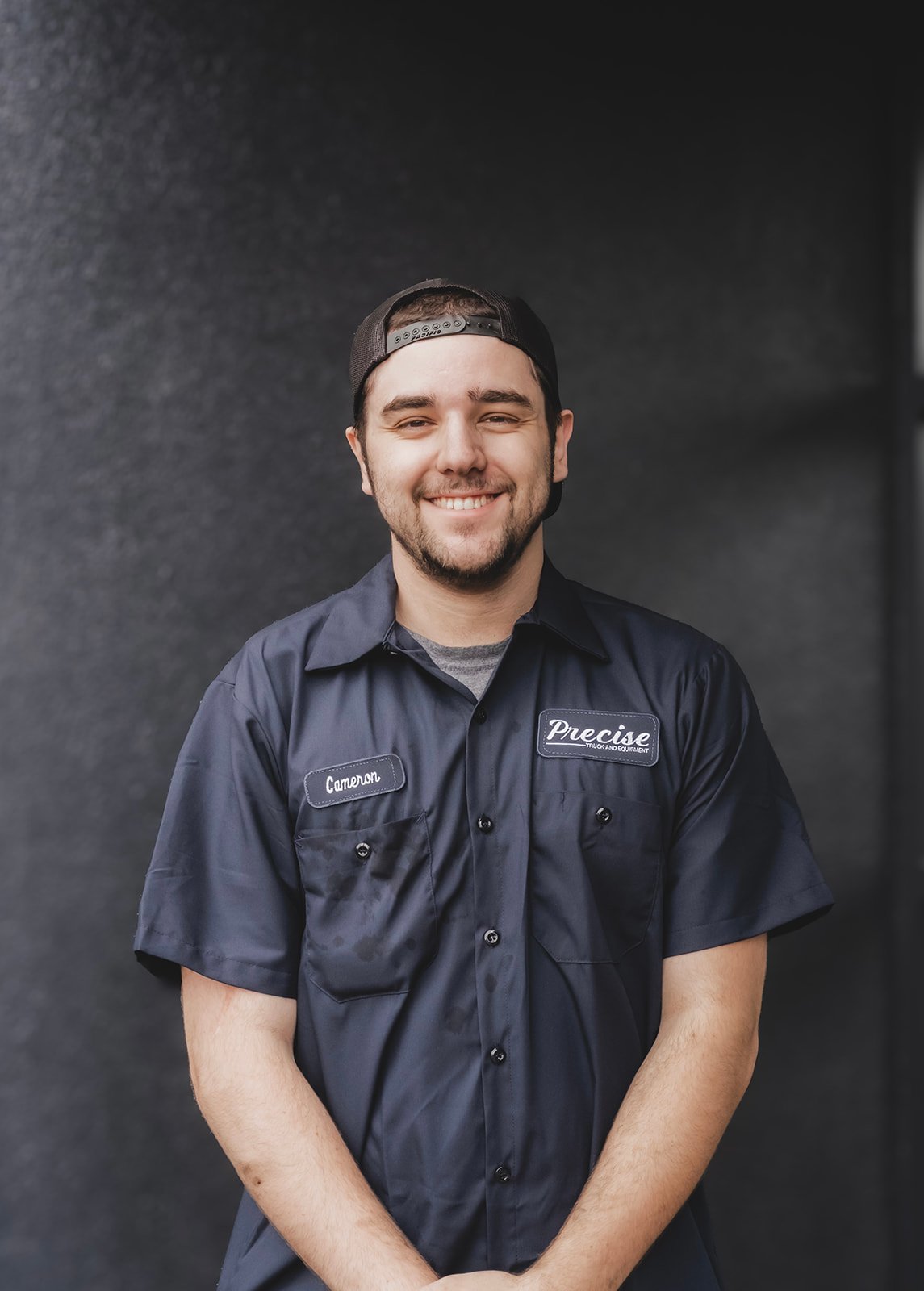 A smiling man wearing a navy work shirt with a name tag that says Cameron and a patch that says 'Precise,' standing against a dark gray background.