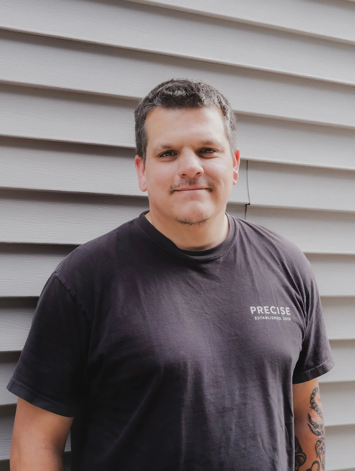 A smiling mechanic wearing a navy blue work uniform standing in a garage. The uniform has a name patch that reads 'Dylan' and a logo patch that says 'Precise.' In the background, there are organized shelves with tools, spray bottles, and a whiteboard on the wall.