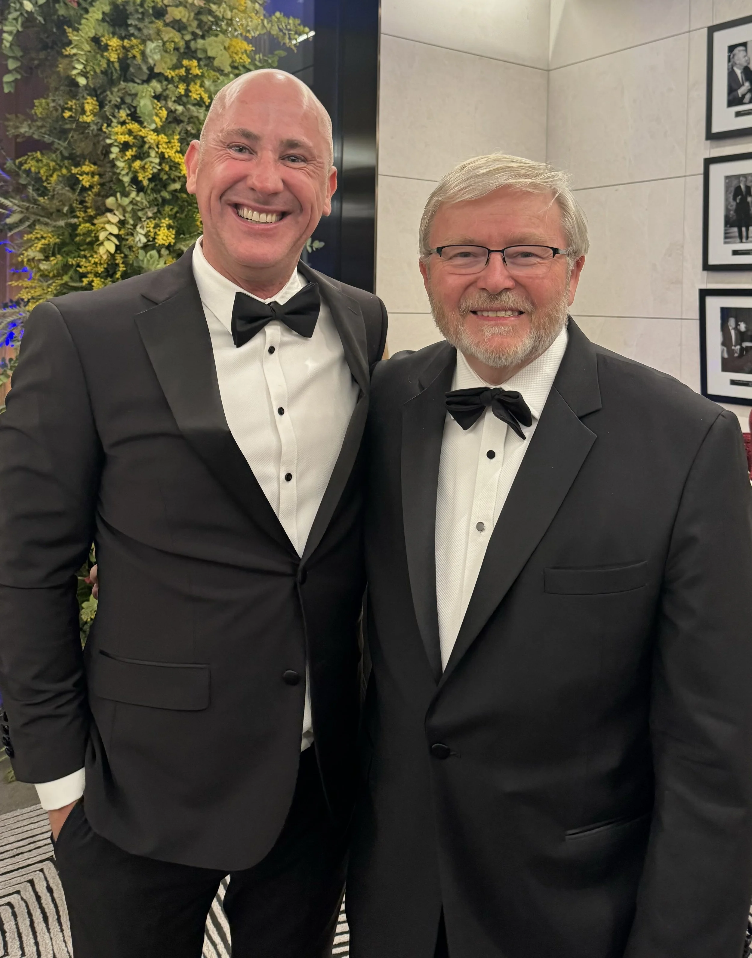 Australian Ambassador Kevin Rudd and Shayne De la Force dressed in black tuxedos with bow ties, smiling at a formal event in an elegant setting with framed black and white photographs on the wall and greenery in the background.