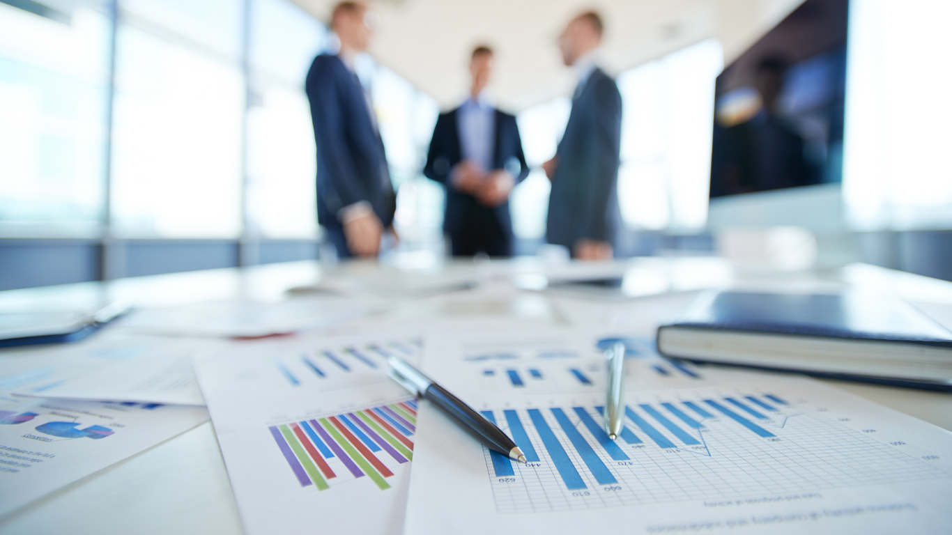 Business meeting with three people in suits in a conference room, and documents with charts on a table in the foreground.