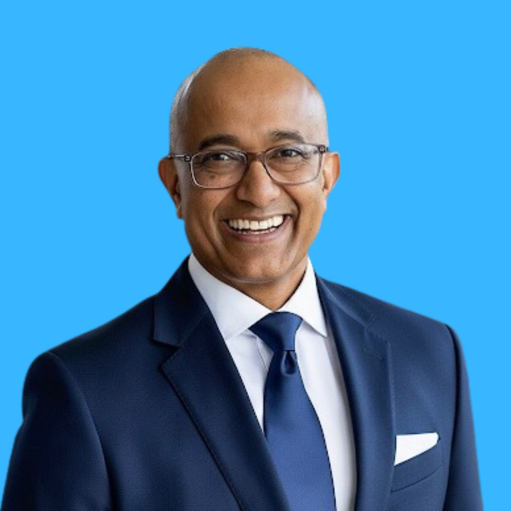 Headshot of a man with a bald head, glasses, and a big smile, dressed in a navy blue suit, white shirt, and blue tie, against a bright blue background.
