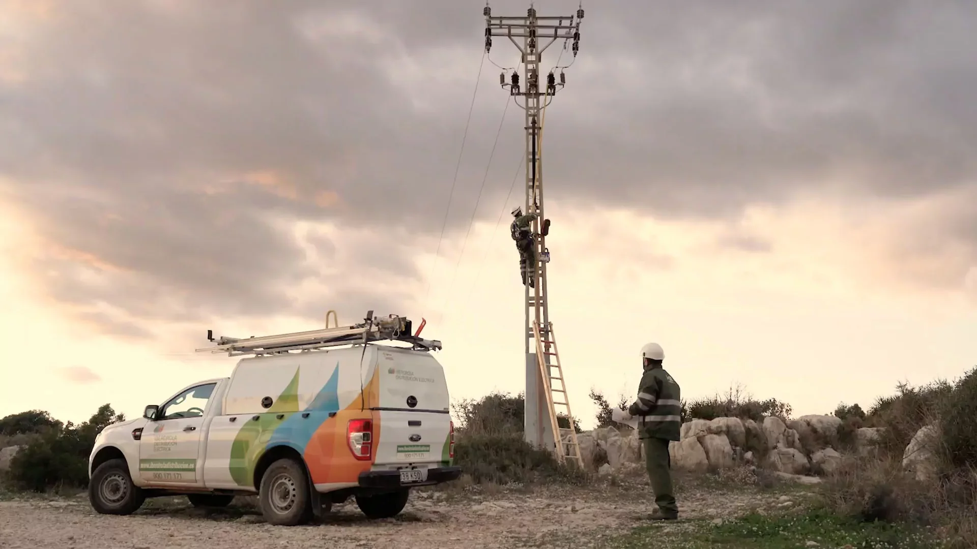 A technician climbing an electrical pole with a safety harness as a worker observes nearby. A service vehicle with colorful branding is parked next to the pole under a cloudy sky.