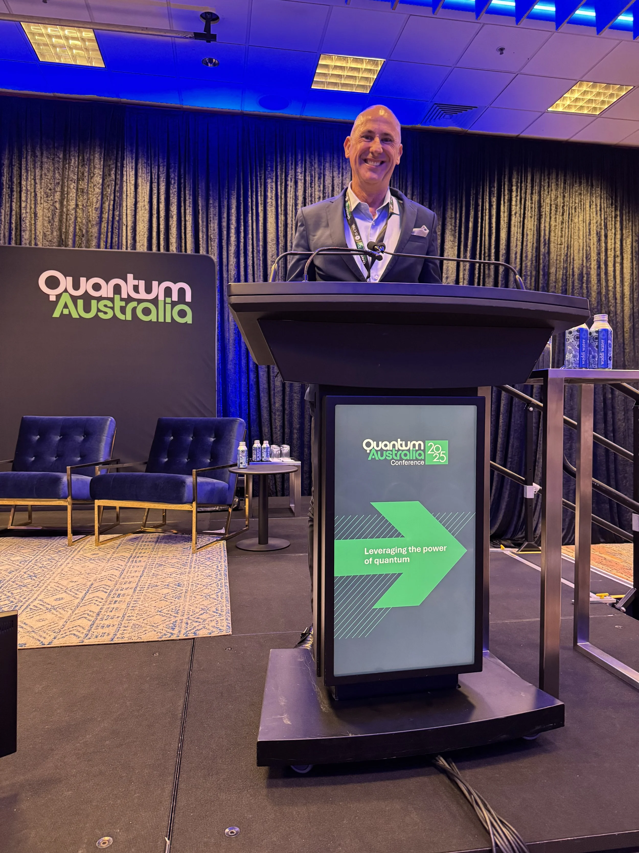 A man in a suit standing at a podium with a screen that reads 'Leveraging the power of quantum' at the Quantum Australia conference 2025, with a stage backdrop and chairs in the background.