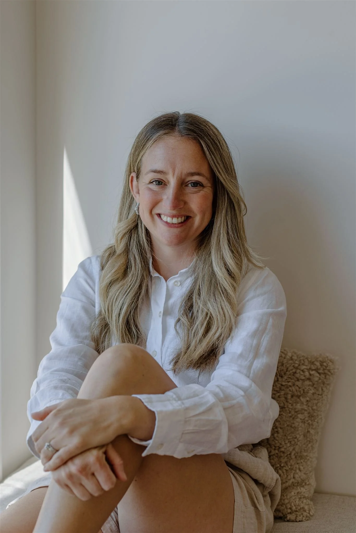 A smiling woman with long, wavy blonde hair working on a laptop at a light wood table decorated with a bouquet of pink and purple flowers, in a bright modern office or craft space.