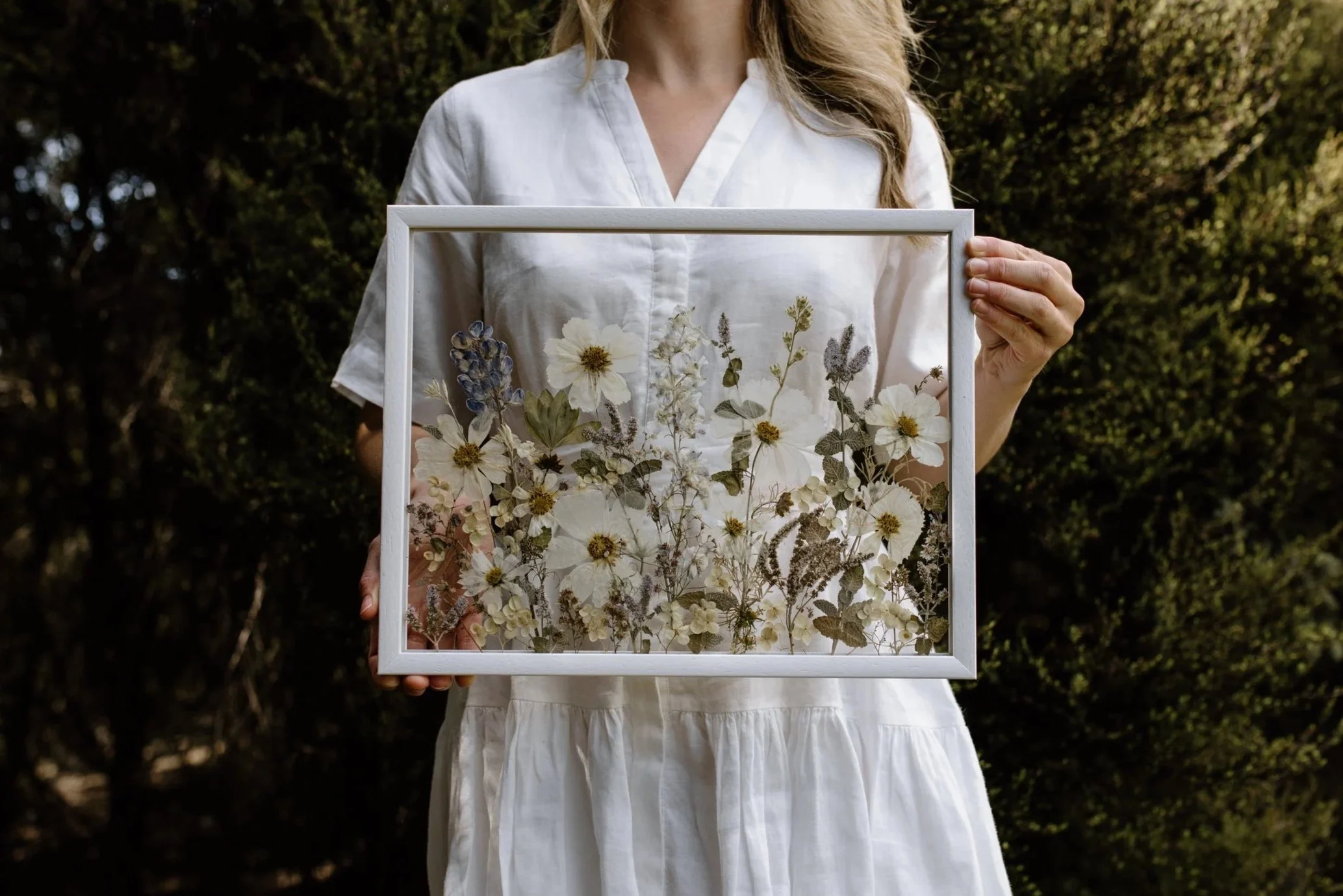 A woman in a white dress holding a framed display of pressed flowers in an outdoor setting.