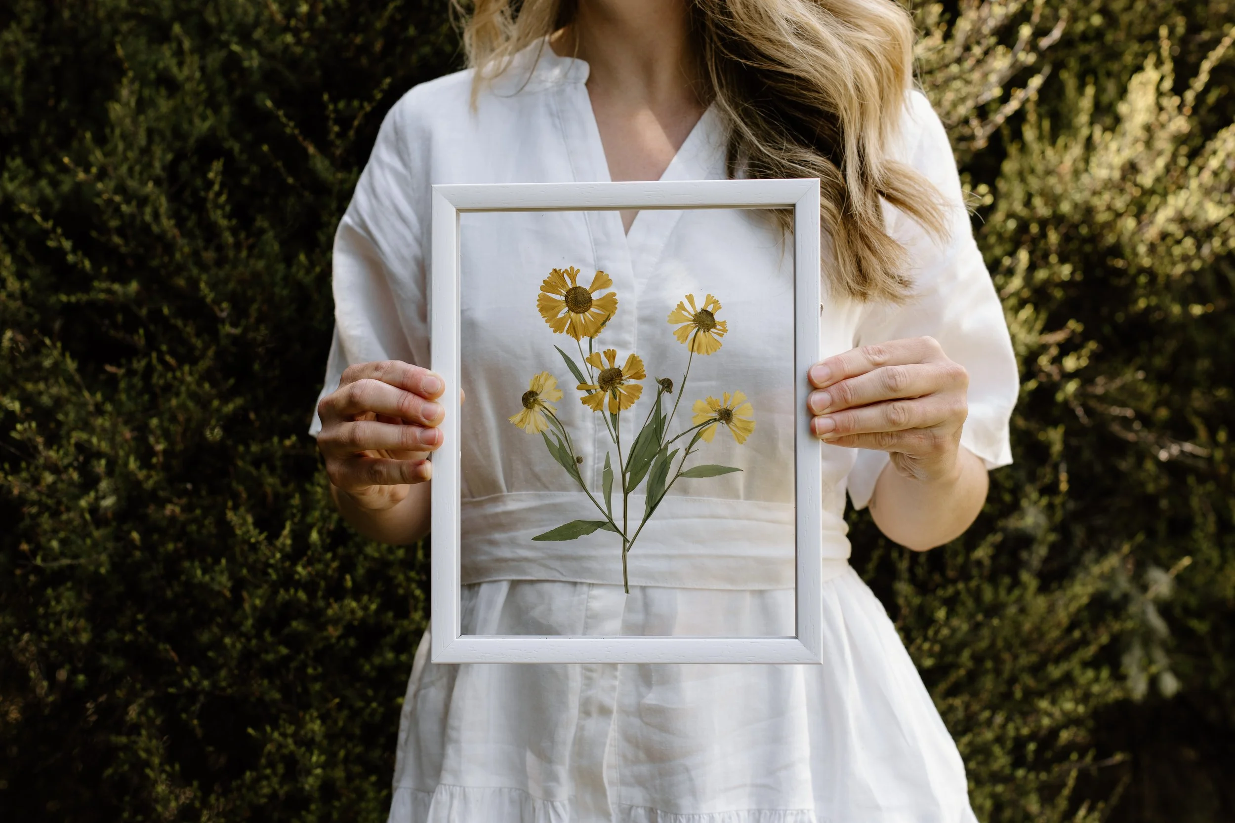 A woman in a white dress holding a picture frame with a pressed yellow flower arrangement inside, standing outdoors with green bushes in the background.