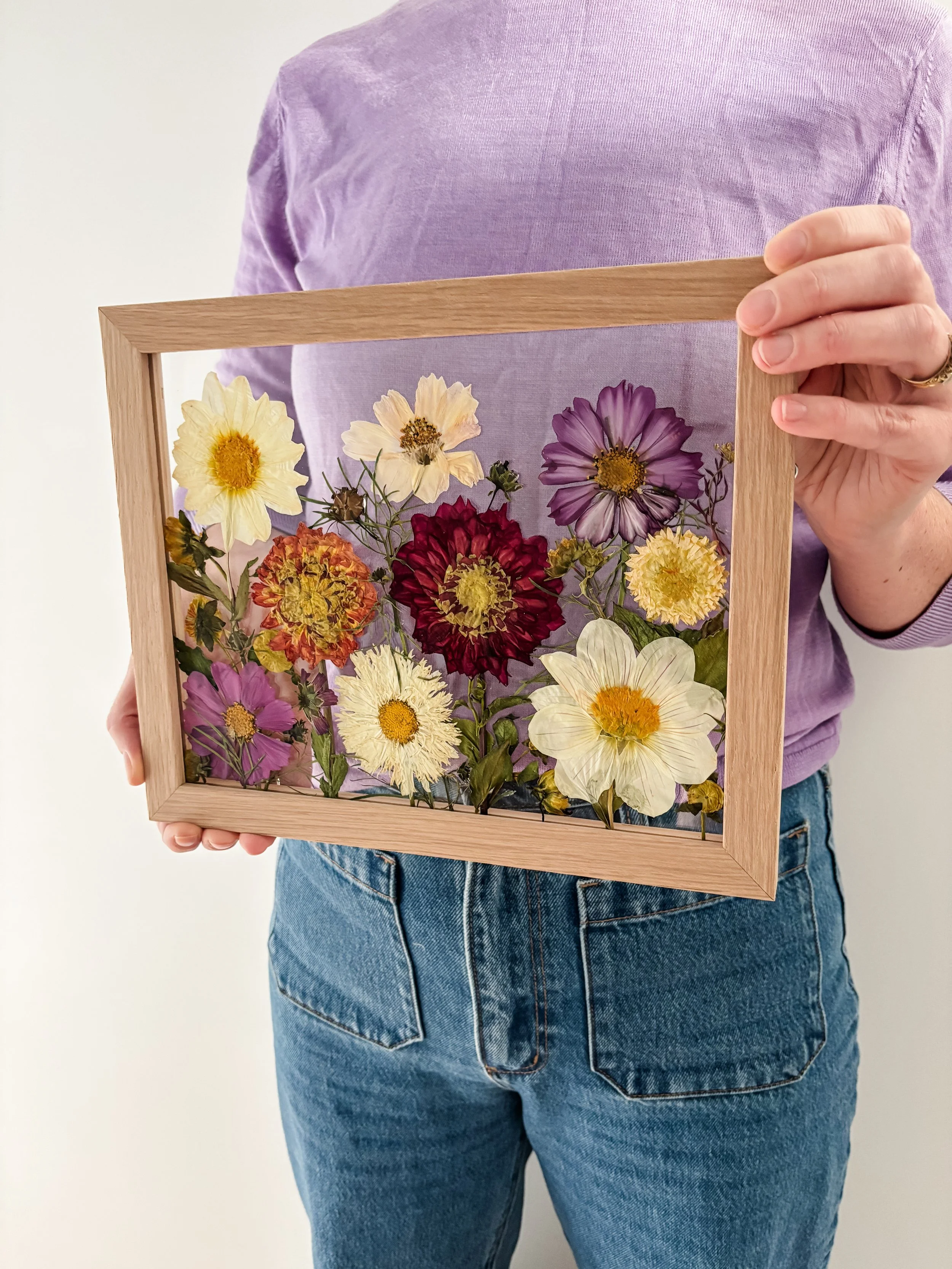 Person holding a wooden frame containing an arrangement of colorful pressed flowers, including yellow, purple, red, and white blossoms.