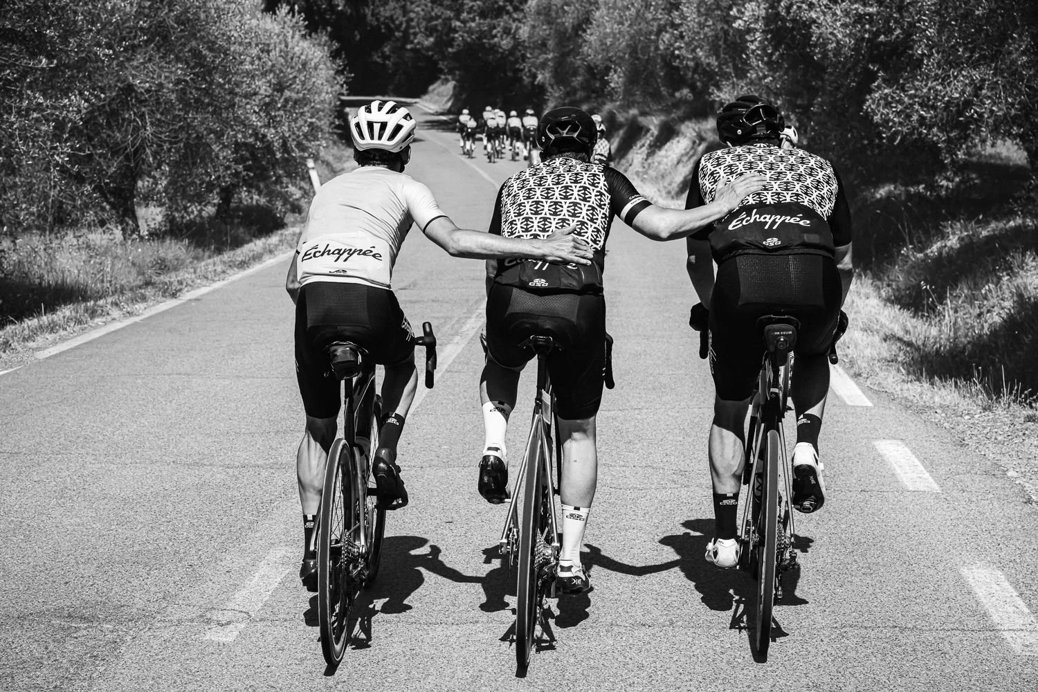 Black and white view from behind of three cyclists riding close together, one with an arm around another's shoulder