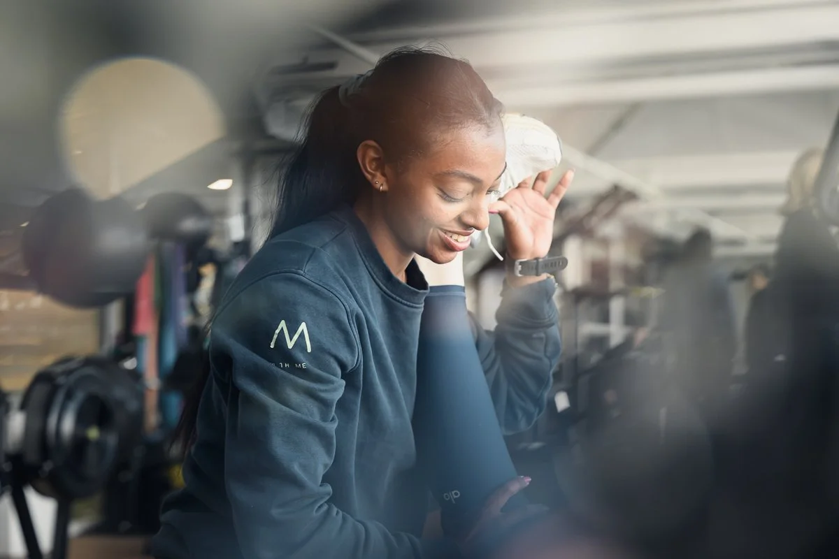 Smiling Fit With Me trainer in a navy sweatshirt looking down, gym equipment soft in the foreground