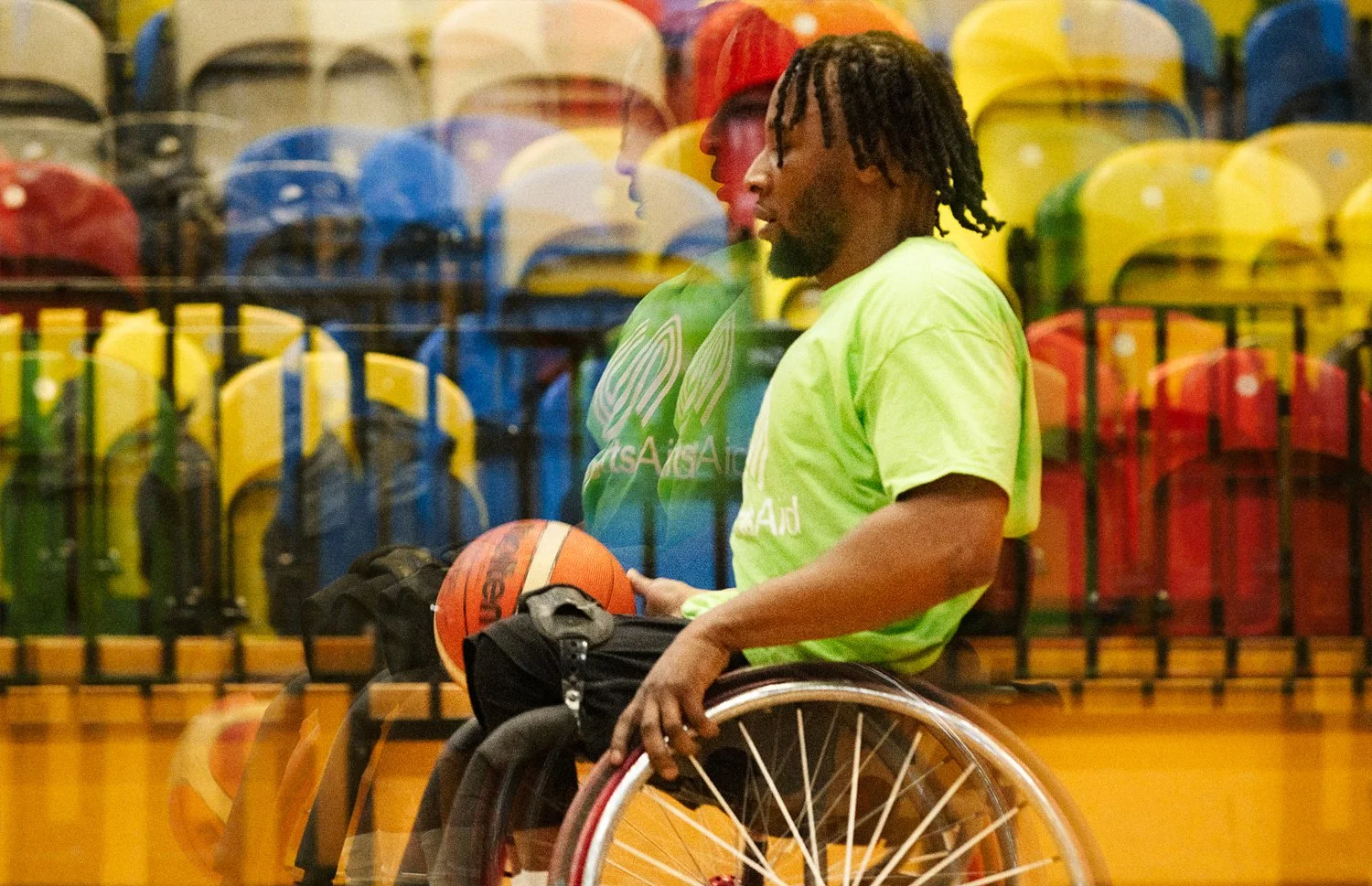 Wheelchair basketball player in a lime green shirt holding a basketball beside the stands, SportsAid