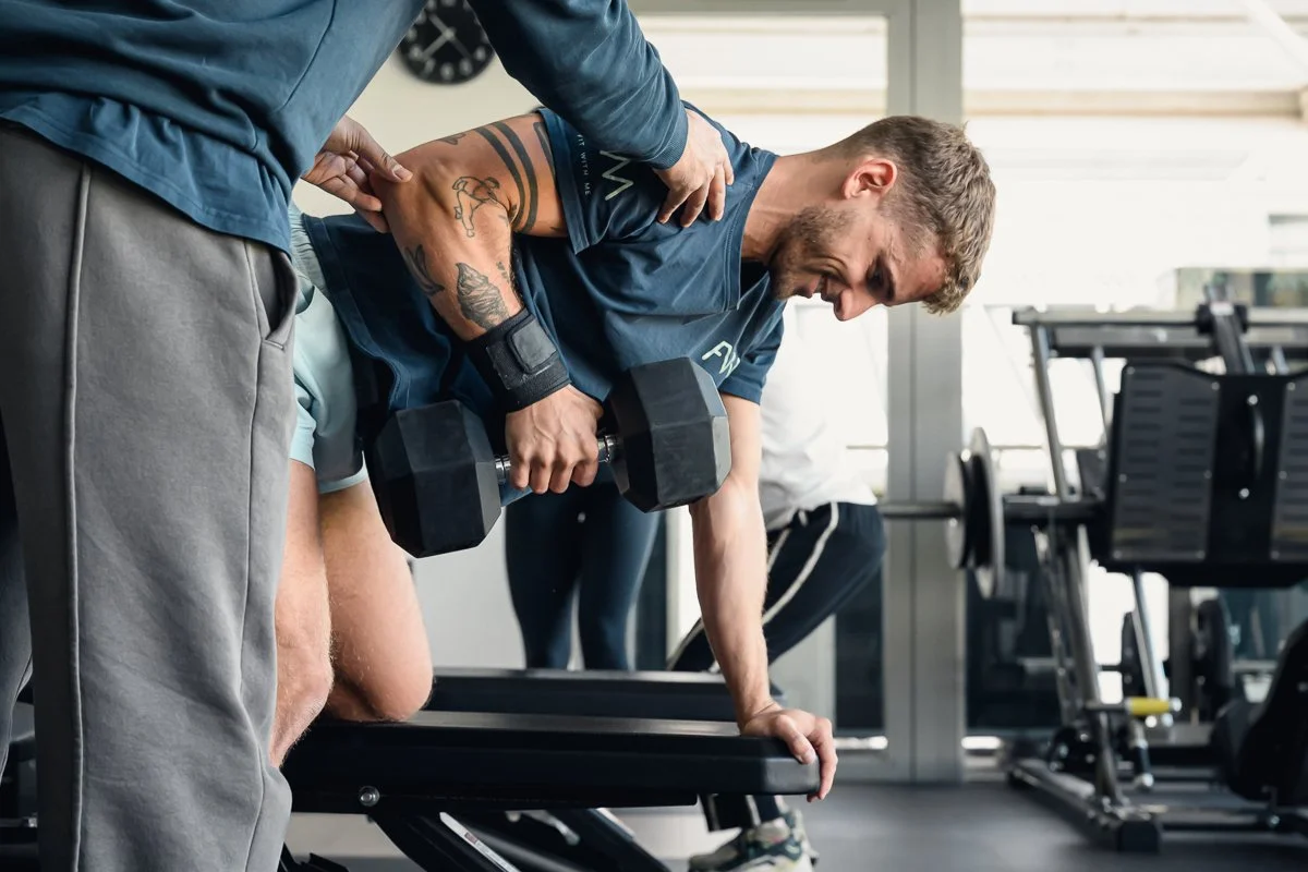 Man smiling through a one arm dumbbell row on a bench while a second person guides his shoulder
