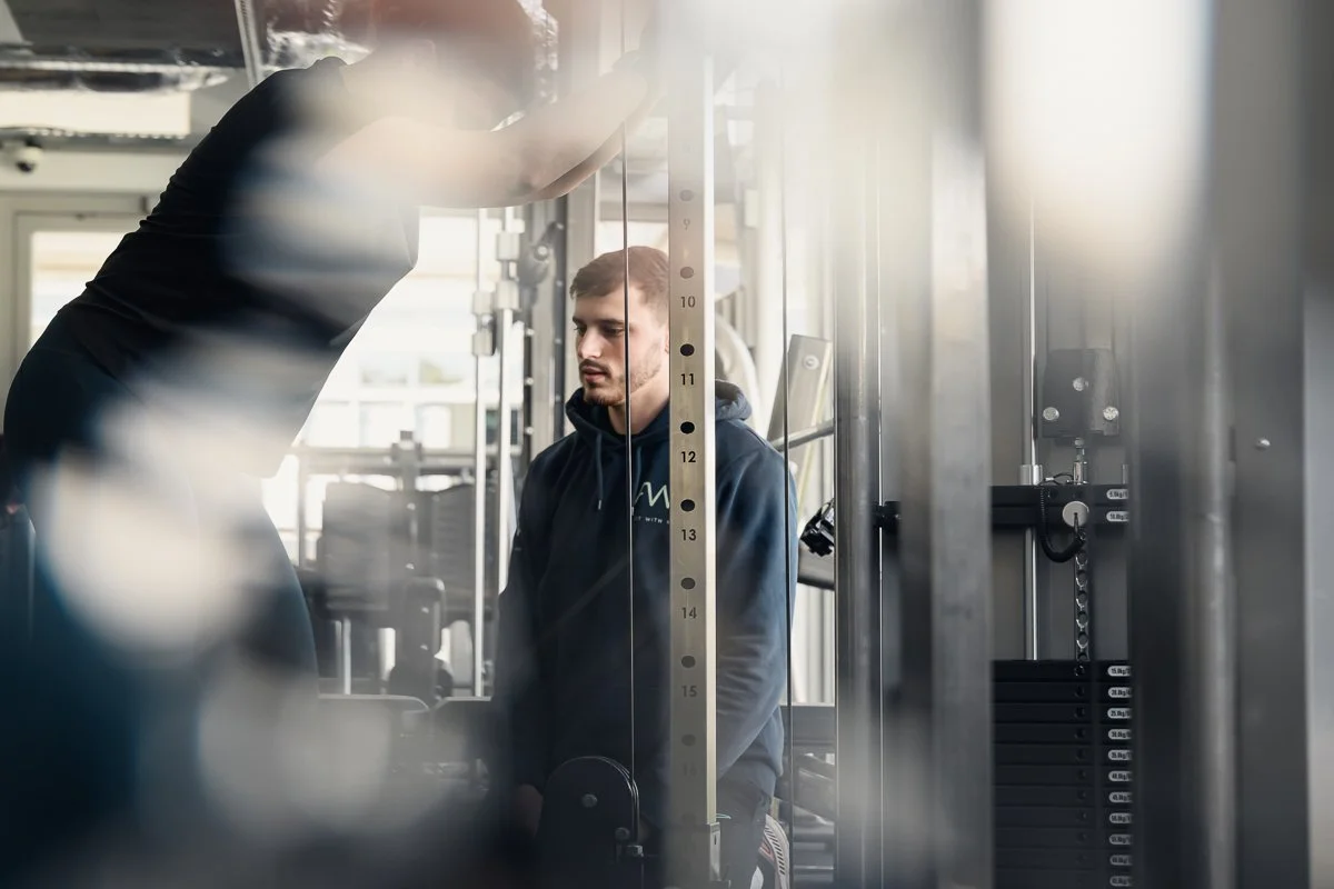 Man in a navy Fit With Me hoodie watching a session from behind a cable station, foreground softly blurred