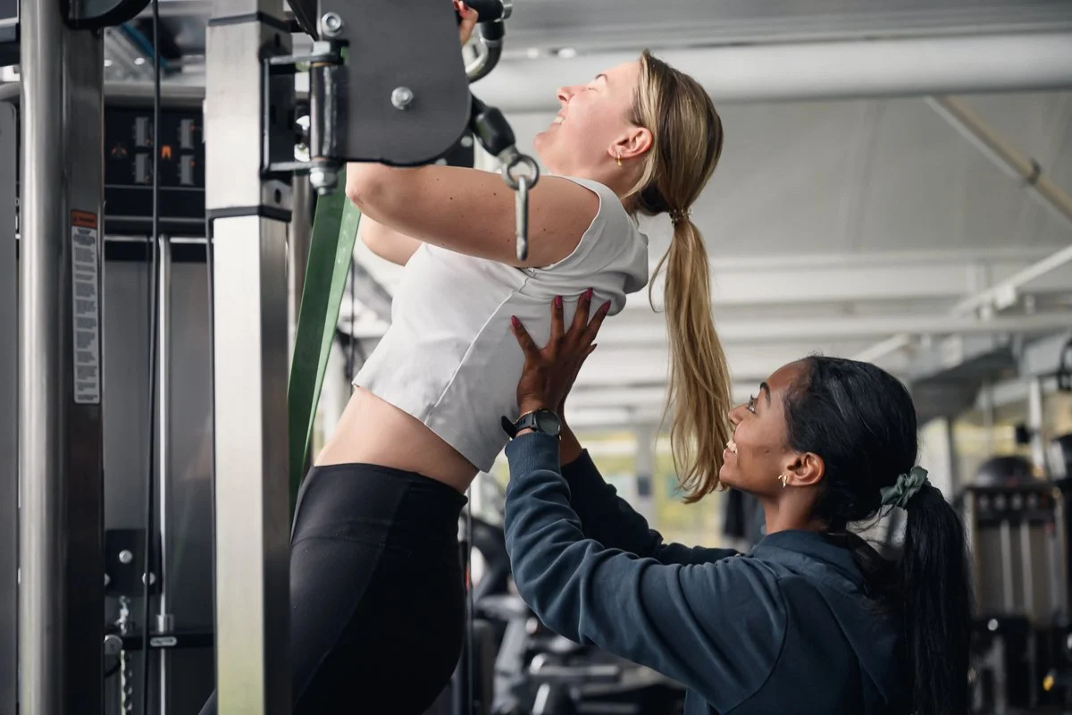 Trainer steadying a laughing client during an assisted pull up at the cable station