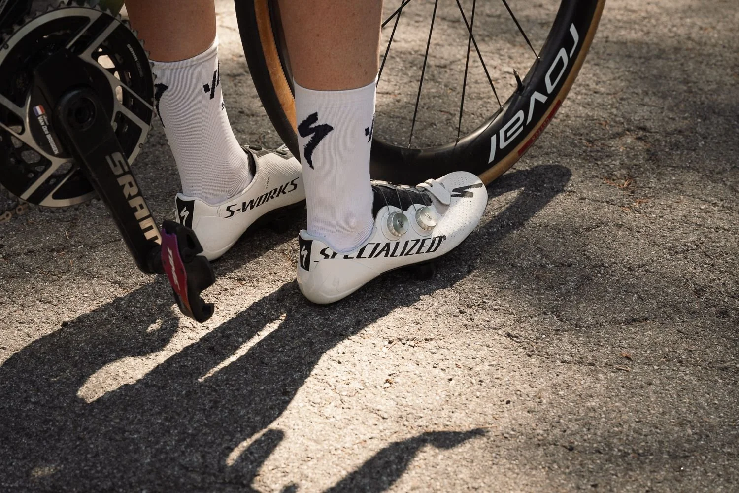 Close detail of white cycling shoes clipped into pedals on a gravel road with long shadows