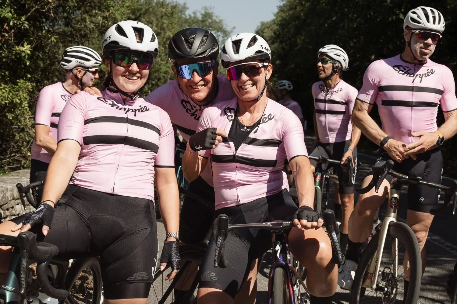 Smiling group of cyclists in pink striped Échappée jerseys standing with their bikes