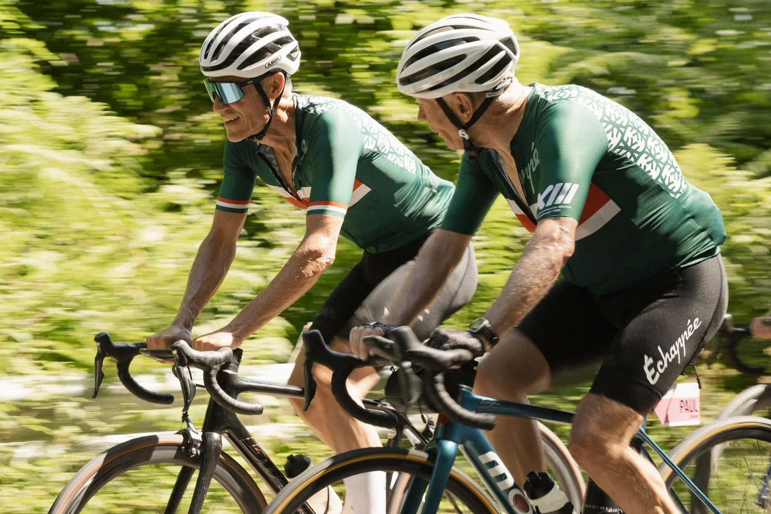 Two cyclists in green jerseys riding side by side, one laughing, with blurred greenery behind