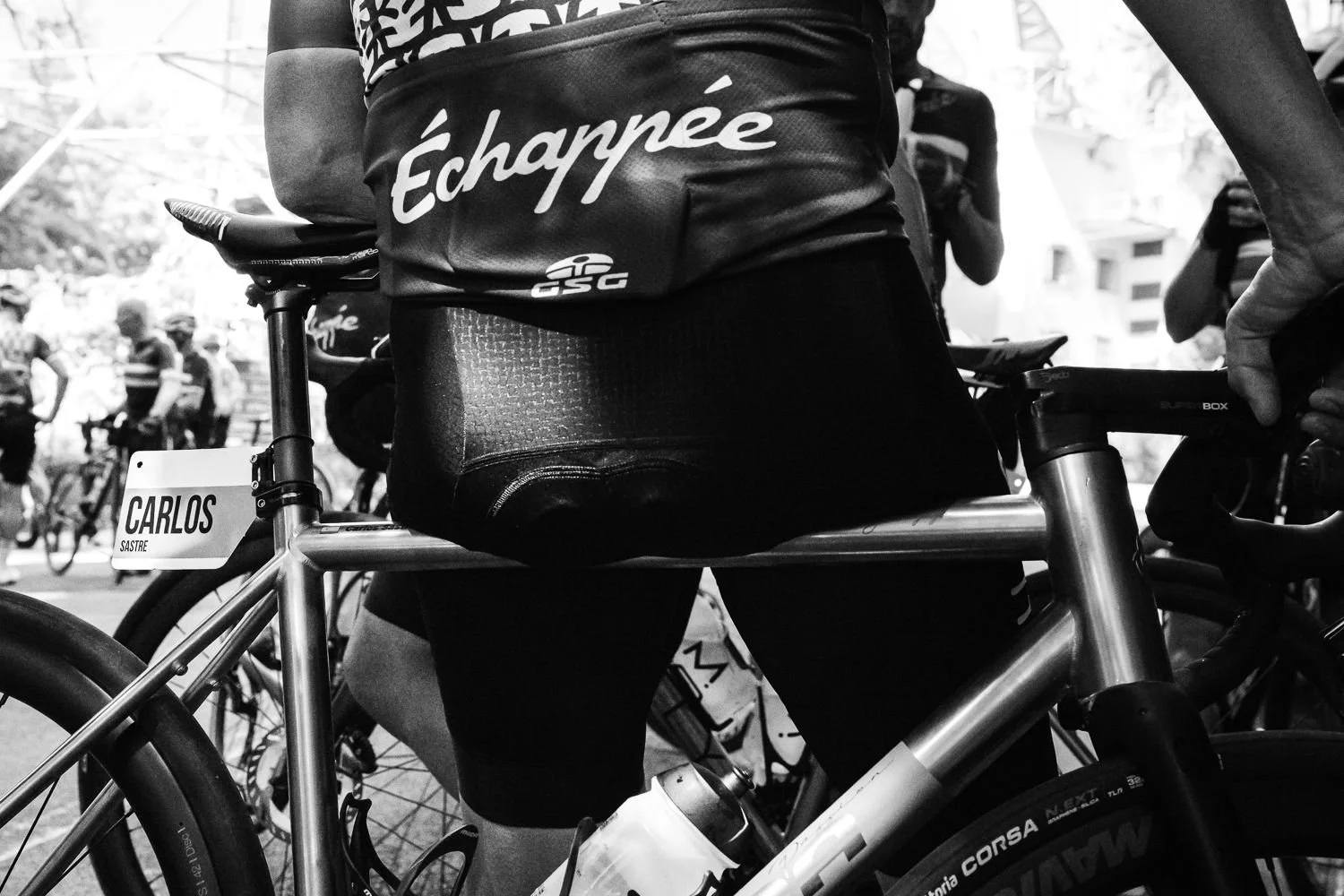 Black and white detail of an Échappée cycling jersey and saddle with riders behind