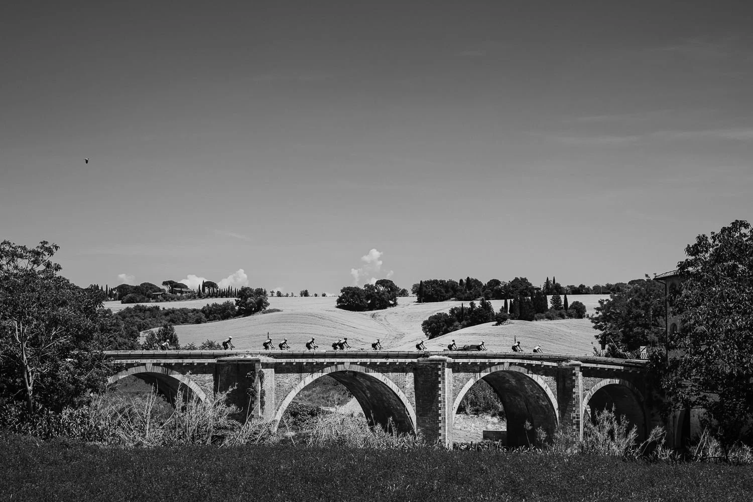 Black and white view of a line of small riders crossing a stone arch bridge with Tuscan hills behind
