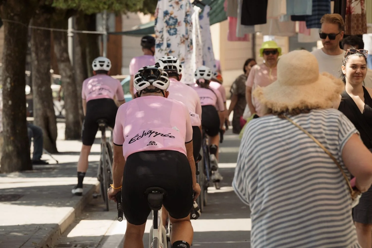 Cyclists in pink jerseys riding slowly through a Tuscan village street past pedestrians