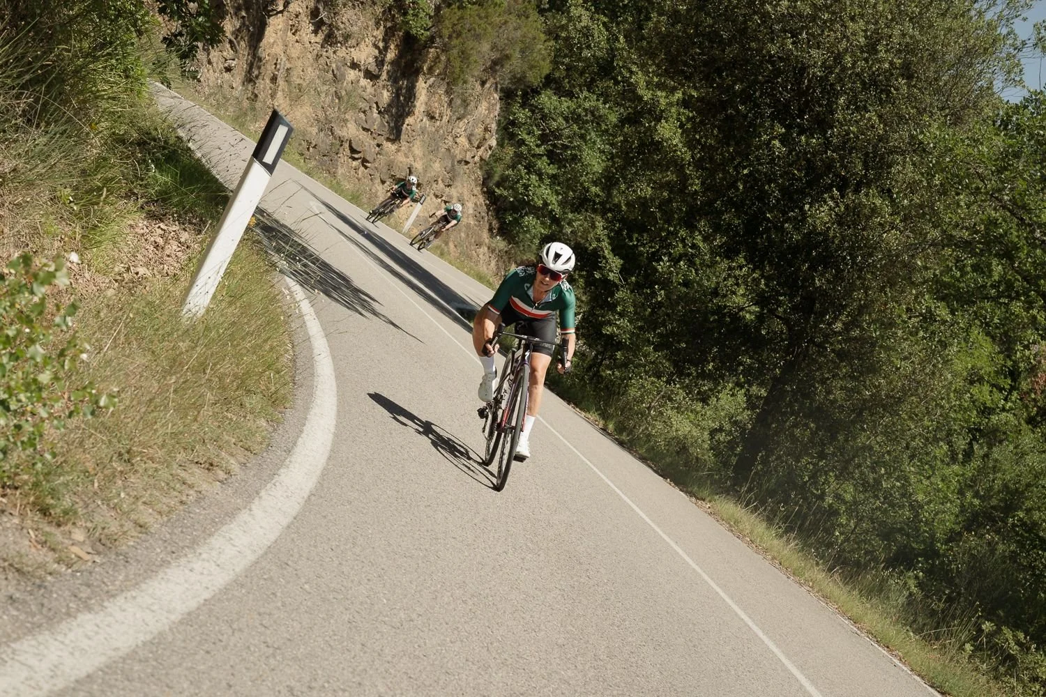 Lone cyclist descending a curving Tuscan road beside a rock face with riders behind
