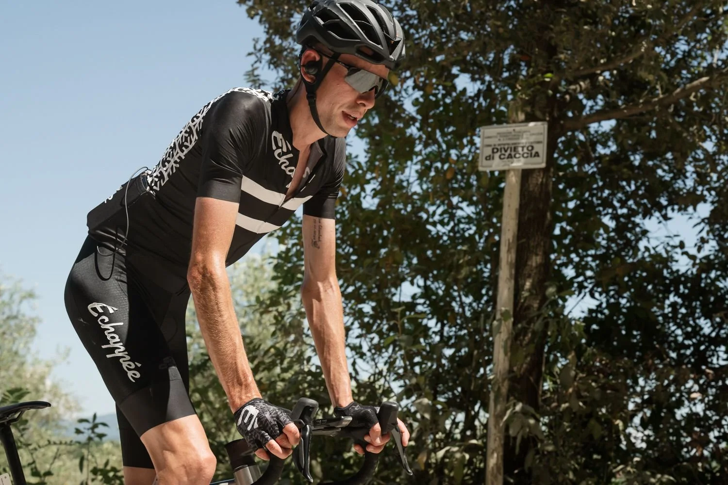 Cyclist in black Échappée kit standing beside a wooden trail sign under trees