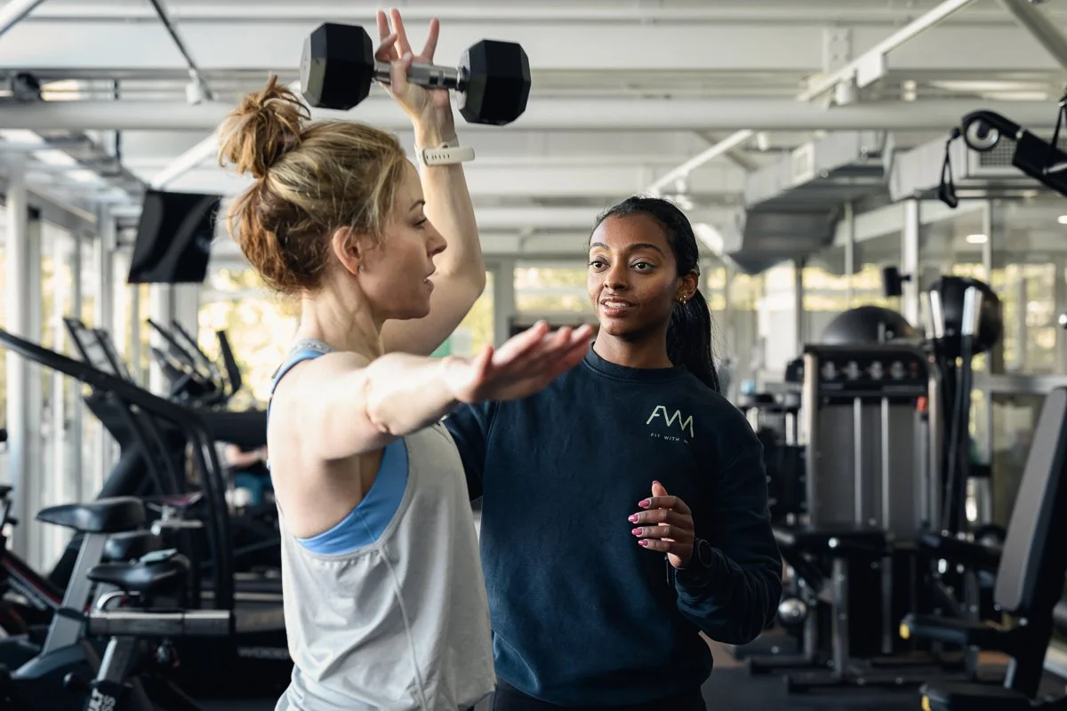 Fit With Me trainer guiding a client through an overhead dumbbell press in the gym at Lord's