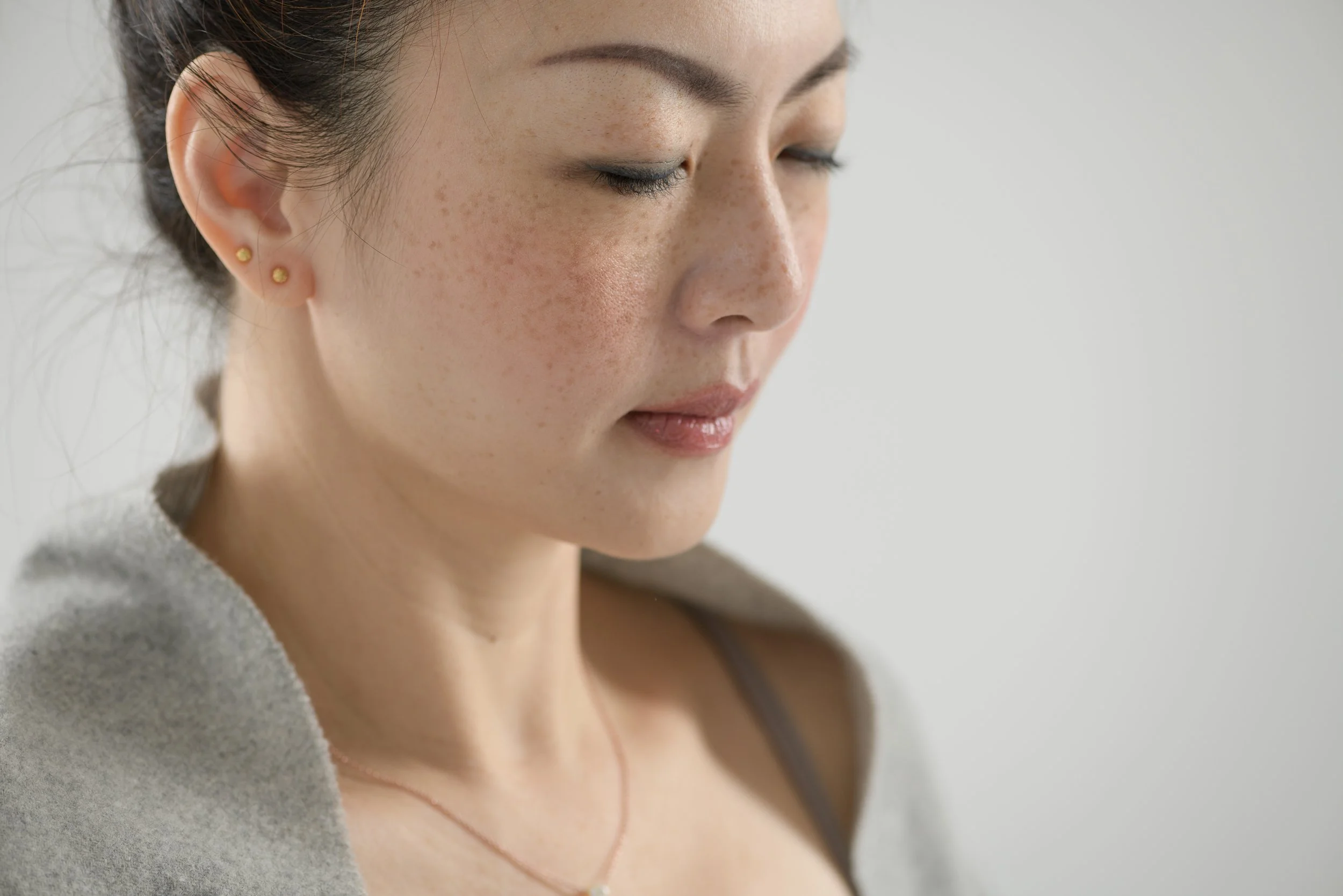 Adelene Cheong in close portrait, eyes closed, wrapped in a soft grey layer, a serene expression against a plain studio backdrop