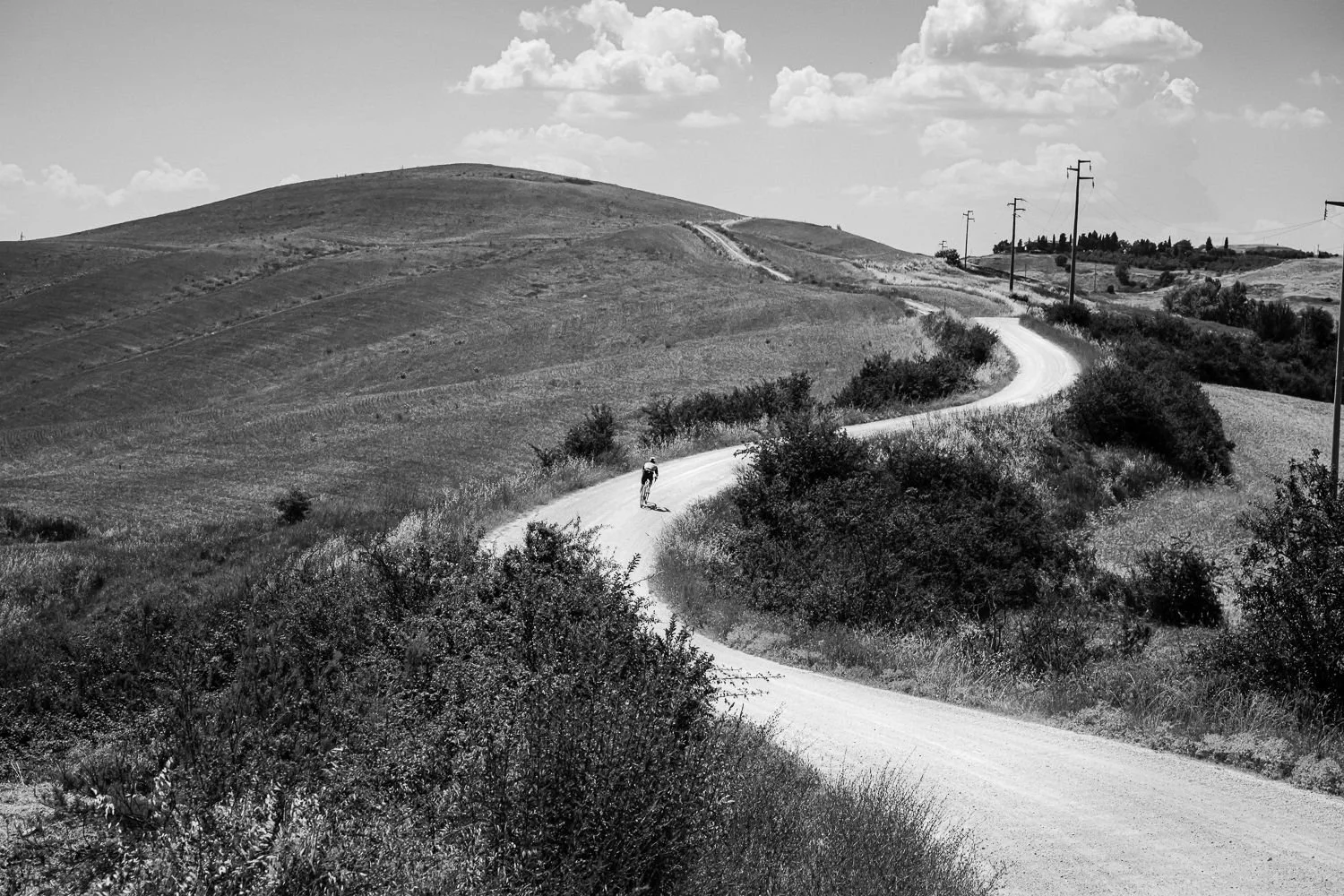 Black and white view of a rider on a winding white gravel road across a rolling hill