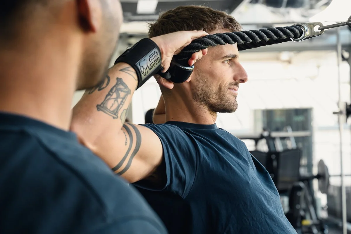Client working a rope cable attachment overhead with a trainer's shoulder in the foreground