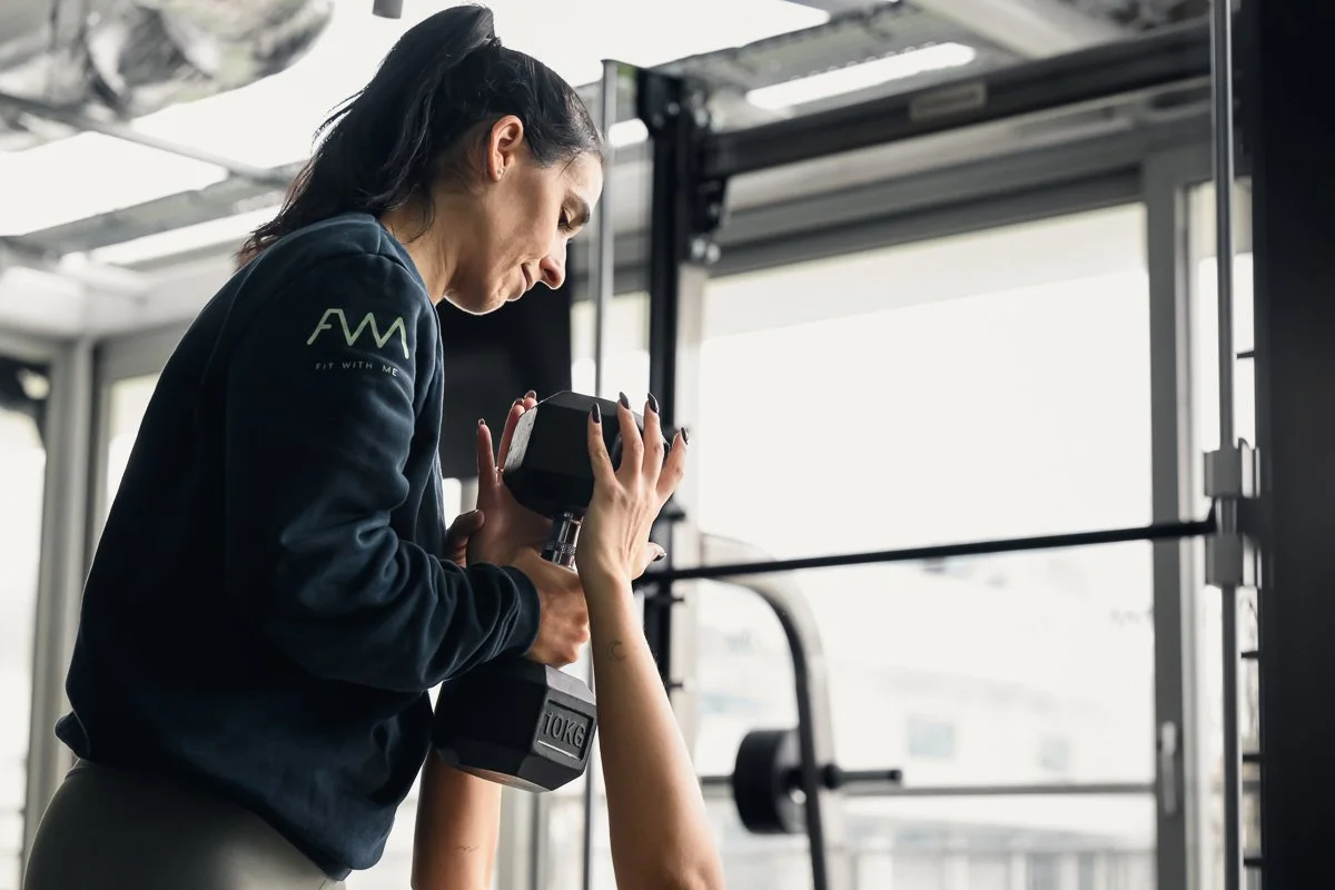 Fit With Me trainer in a navy sweatshirt guiding a client's hands around a 10kg dumbbell, large windows behind her
