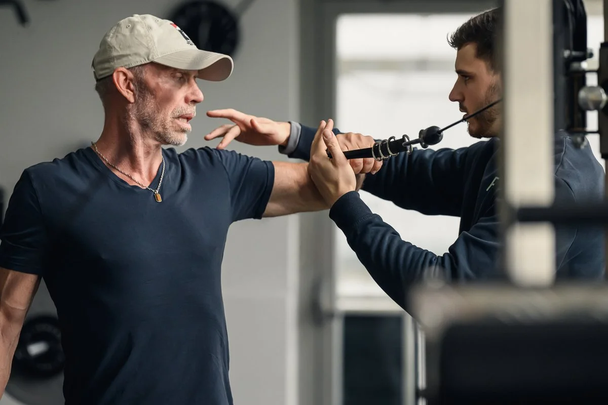 Trainer adjusting a client's arm position at a cable machine, the client in a cap looking on