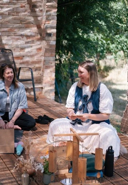 Two women sitting on a wooden deck outdoors, smiling and talking, with one holding a smartphone. There is a brick wall behind them and trees in the background.