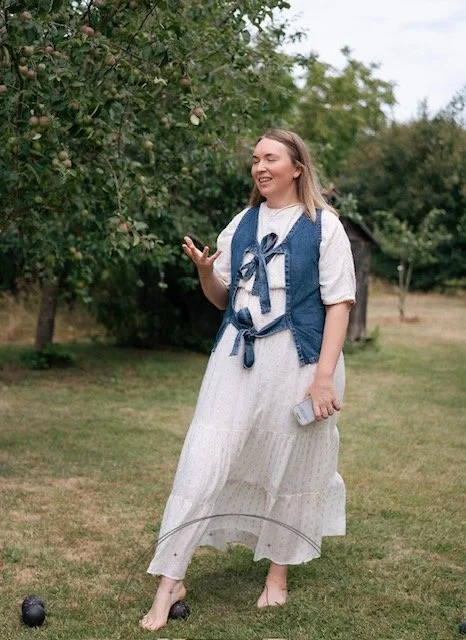 A woman in a white dress and denim vest standing barefoot on a grassy field, holding a water bottle, and reaching towards apple trees with pink apples.
