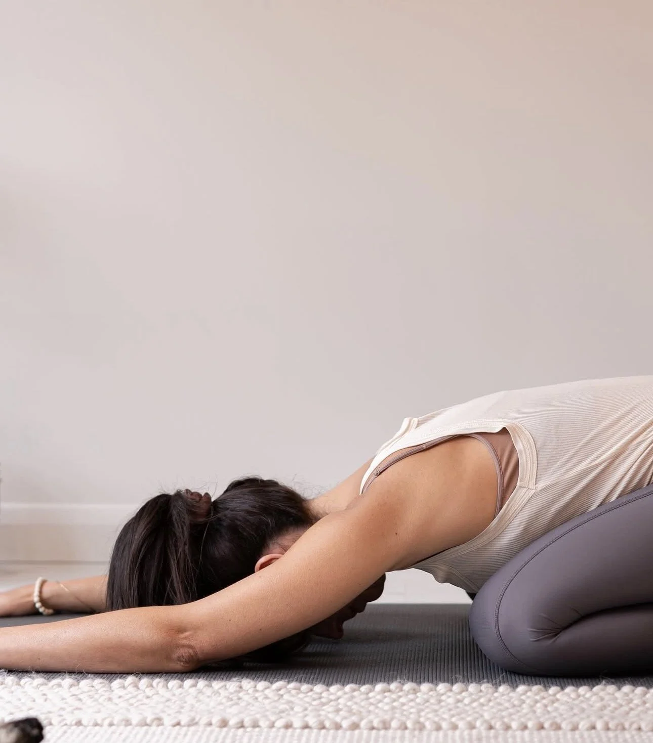 A woman practicing yoga in a child's pose on a yoga mat in a room with beige walls.
