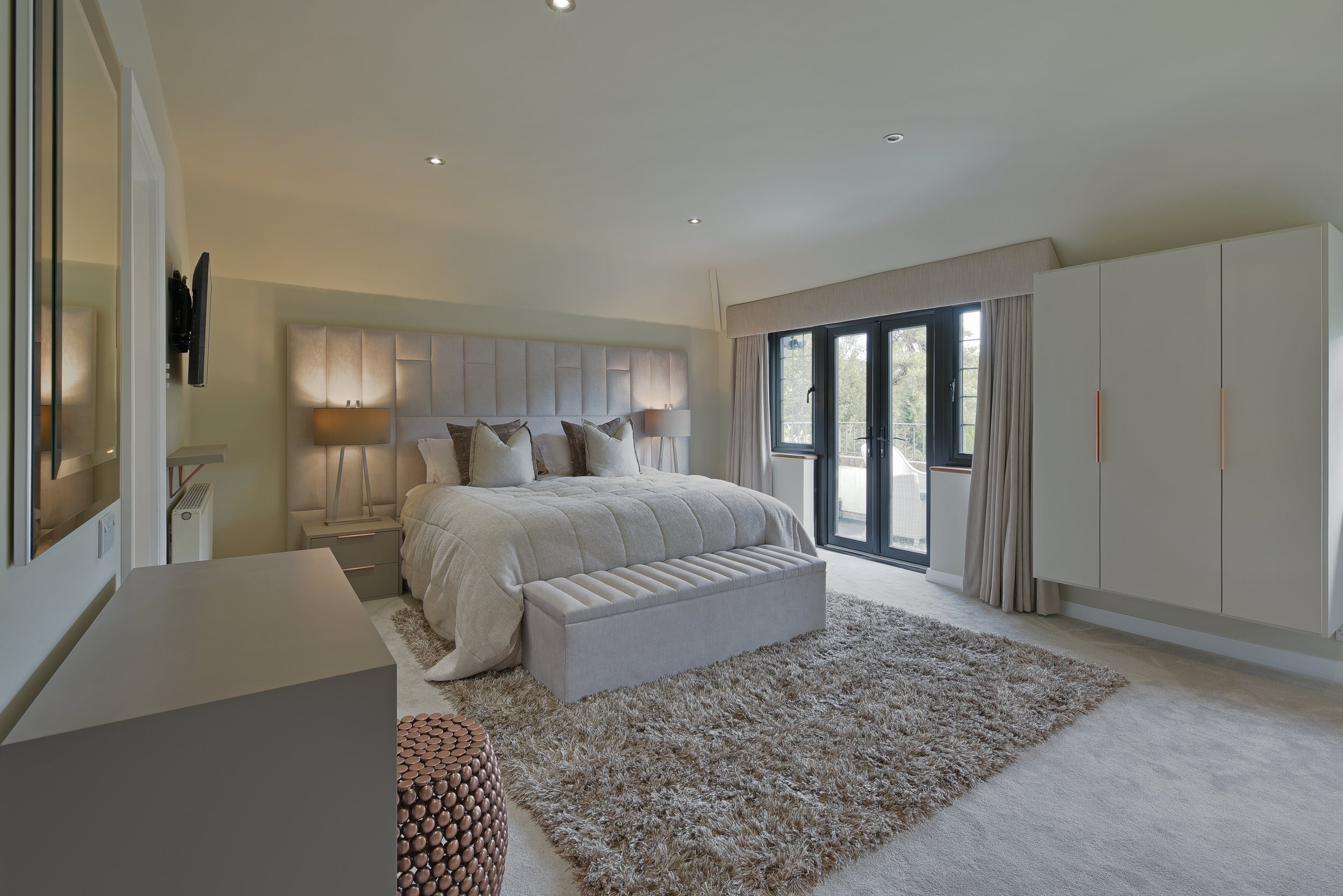 Modern bedroom with large headboard and upholstered bed, bespoke bedside tables, cream curtains, fitted wardrobes.
