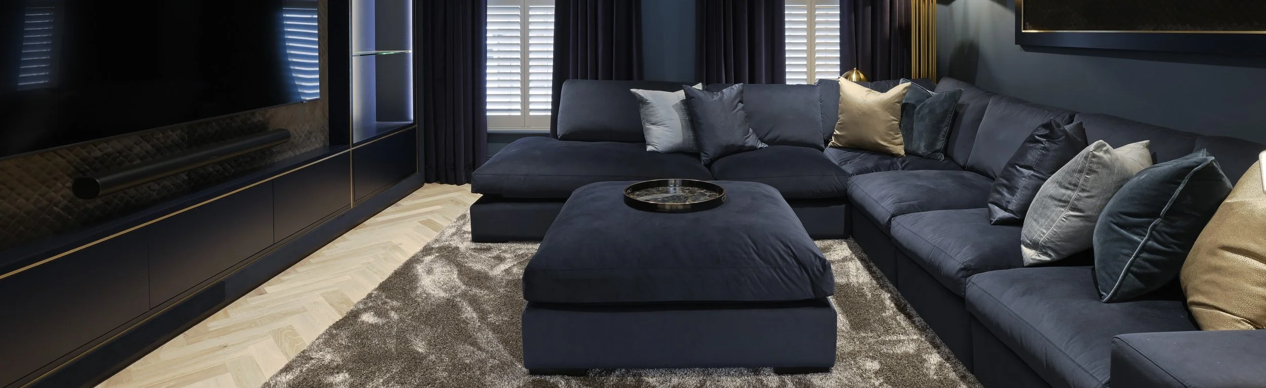 Modern living room with a dark blue sectional sofa, gold and gray pillows, a marble tray on matching blue ottoman, dark curtains, and a large flat-screen TV.