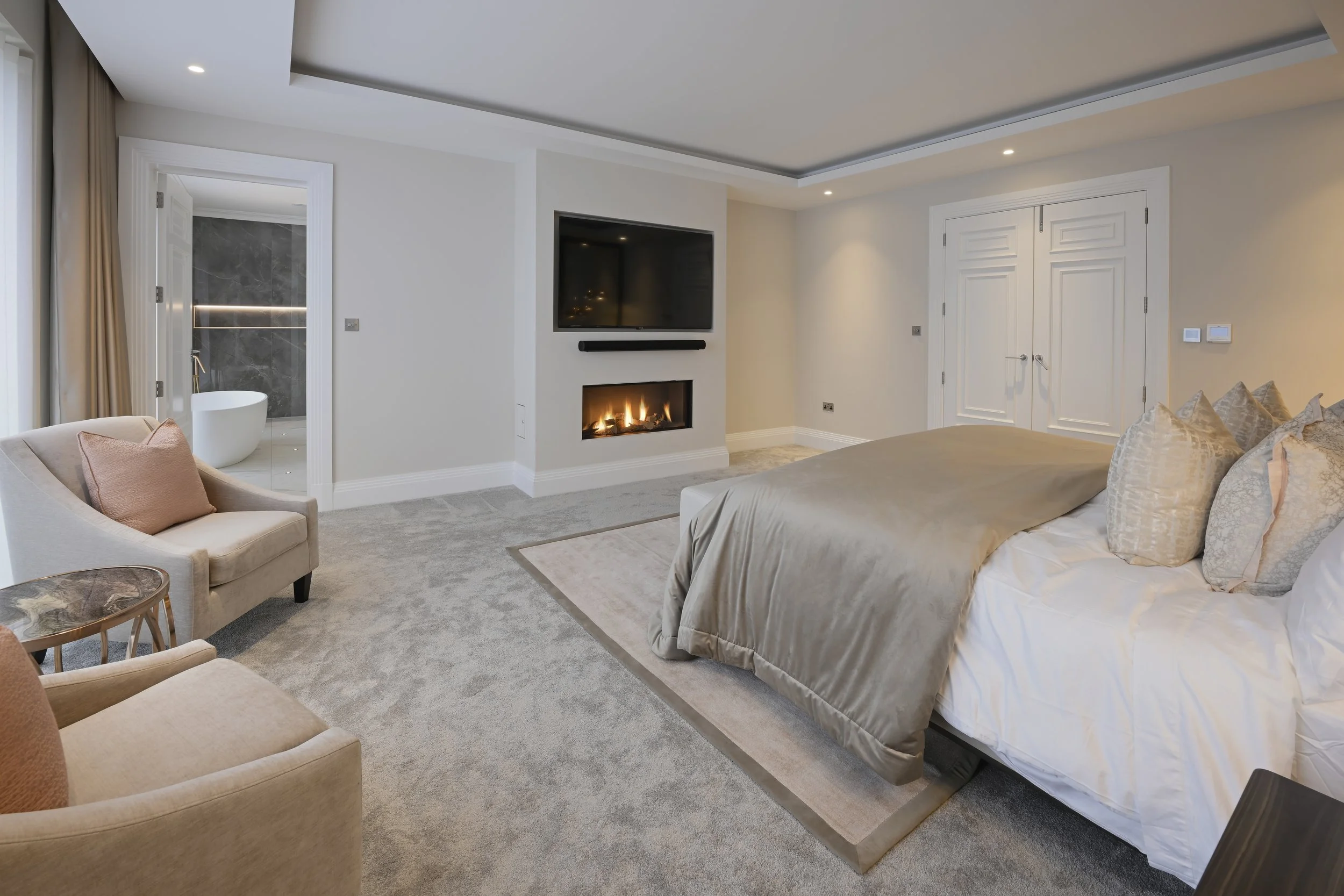 Luxury bedroom with a large bed, fireplace, wall-mounted TV, armchair, and a view into a bathroom with a bathtub.