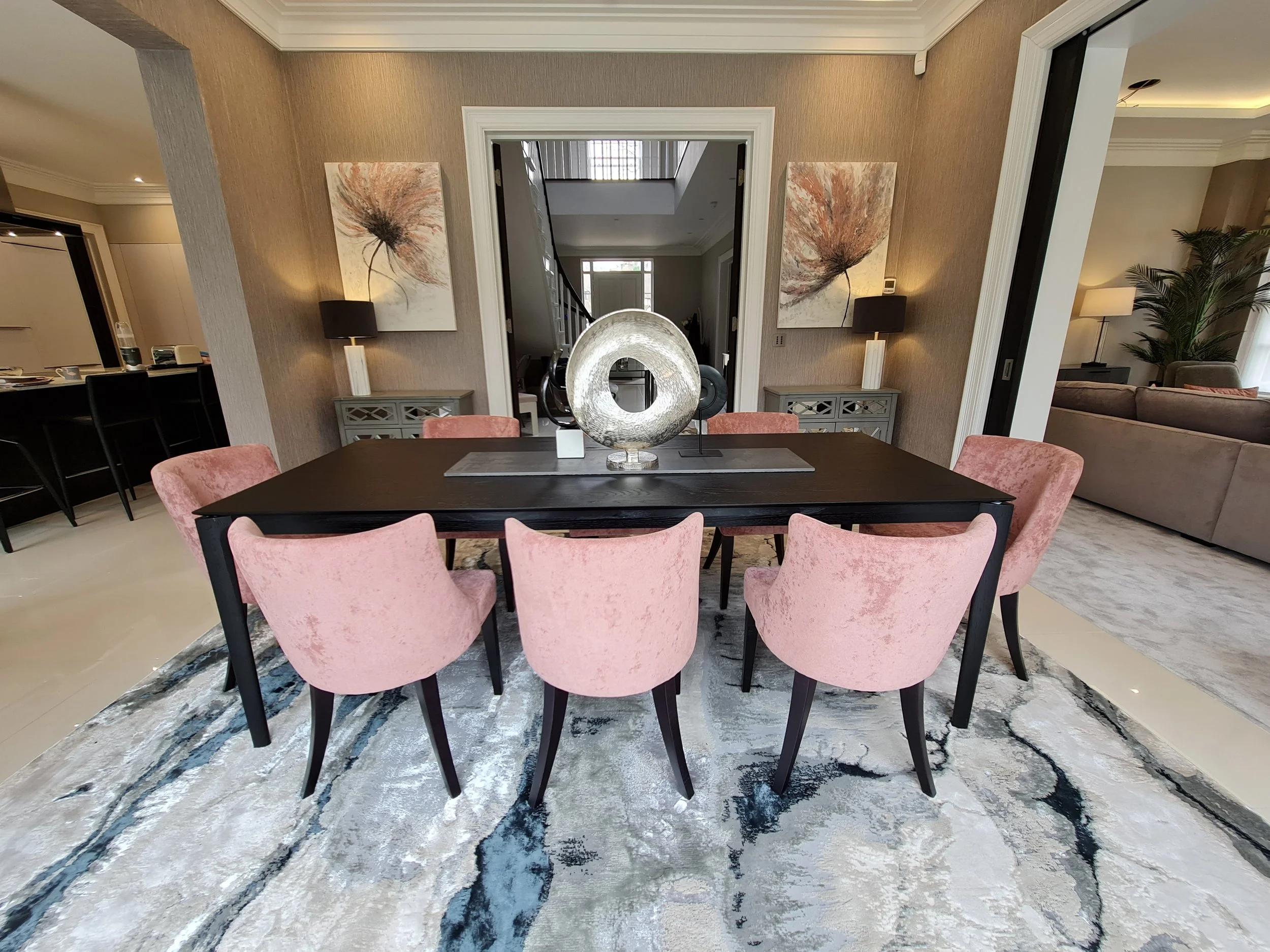 A dining room with a dark oak dining table, pink velvet upholstered chairs, a silver sculpture centerpiece, and modern artwork on the wall, with a living room visible in the background.