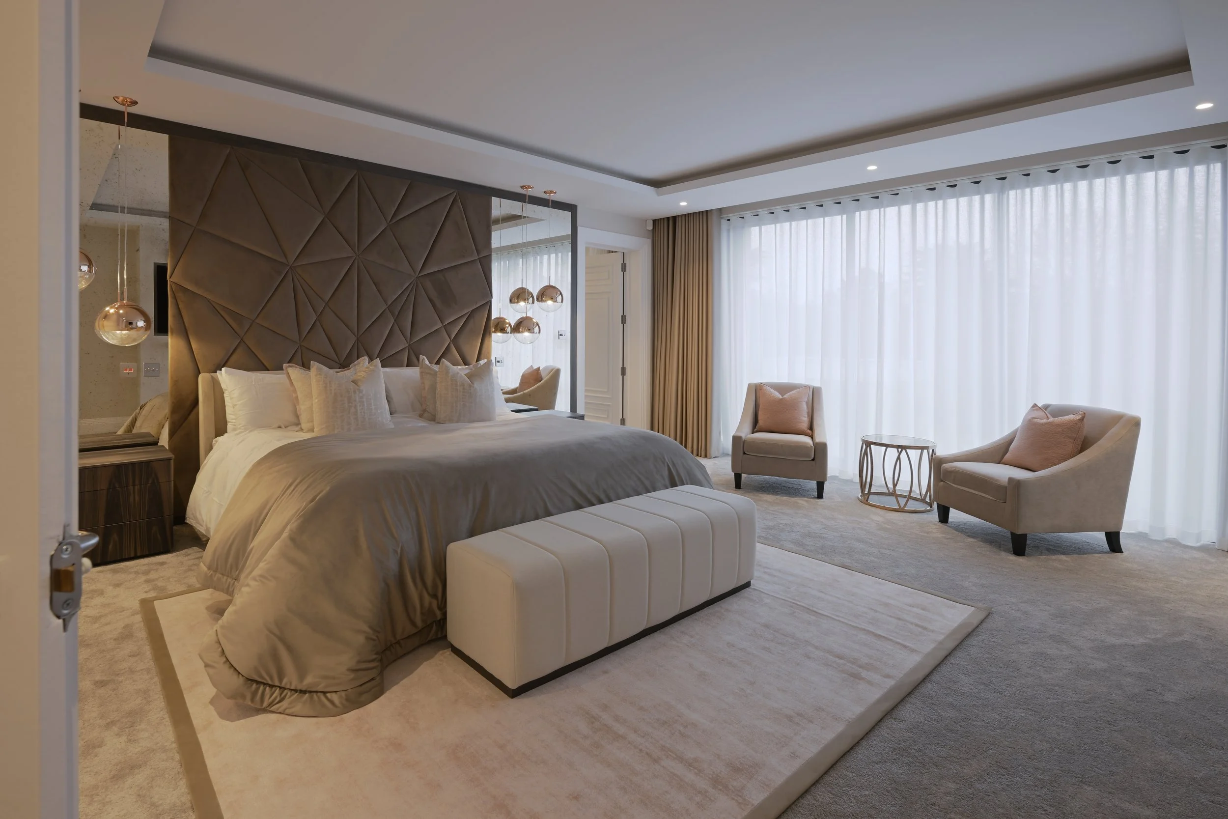 Elegant bedroom with a large bed, velvet bedspread, and multiple pillows, two armchairs with pink pillows, a side table, and sheer curtains covering a large window. coper pendant lights and fitted headboard and mirrors.